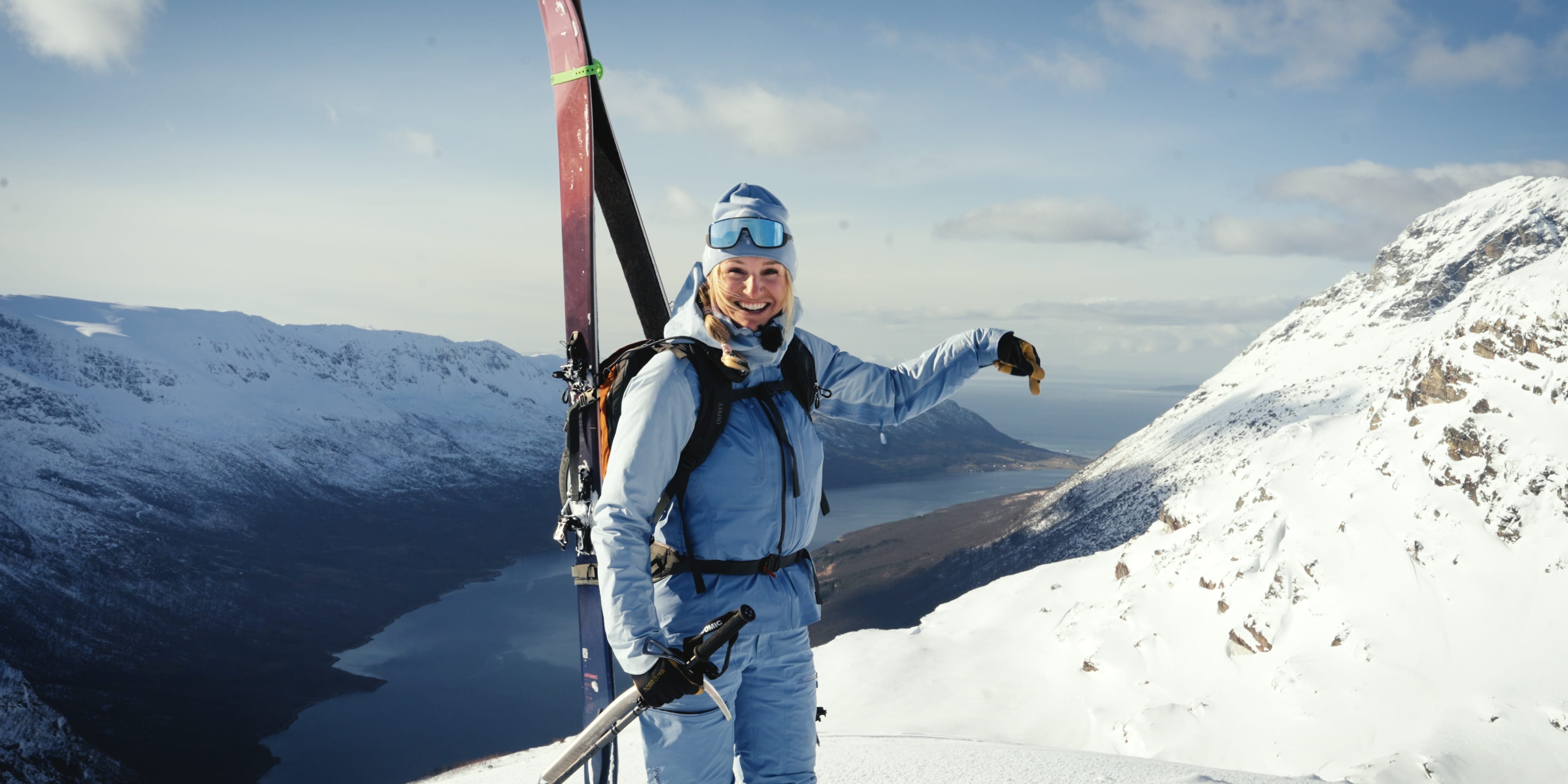 Smiling woman in blue Norrøna ski gear on a snowy mountain peak overlooking a fjord.