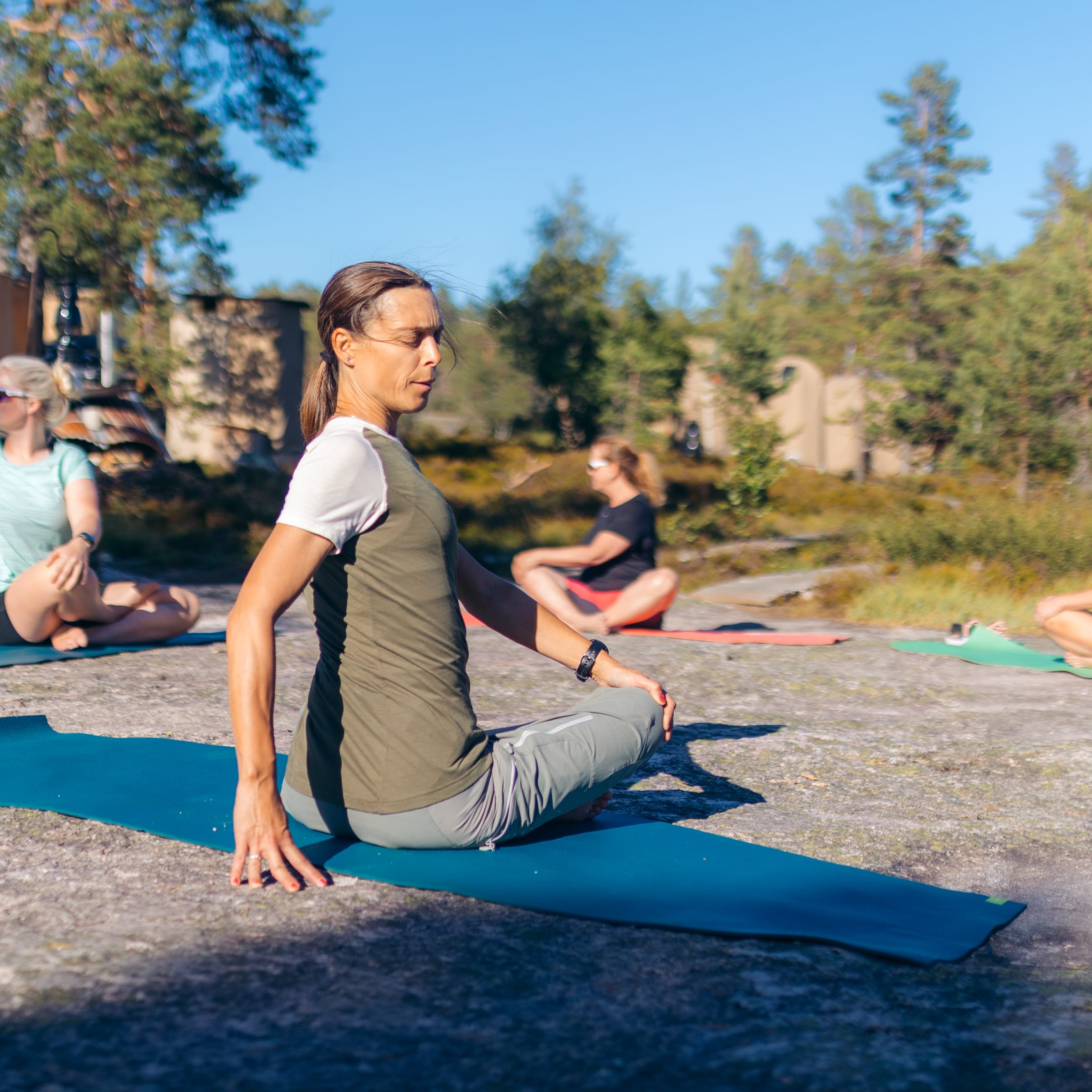 Three people are sitting on yoga mats in a forest clearing. The woman in the foreground is wearing a Norrøna shirt and pants.  
