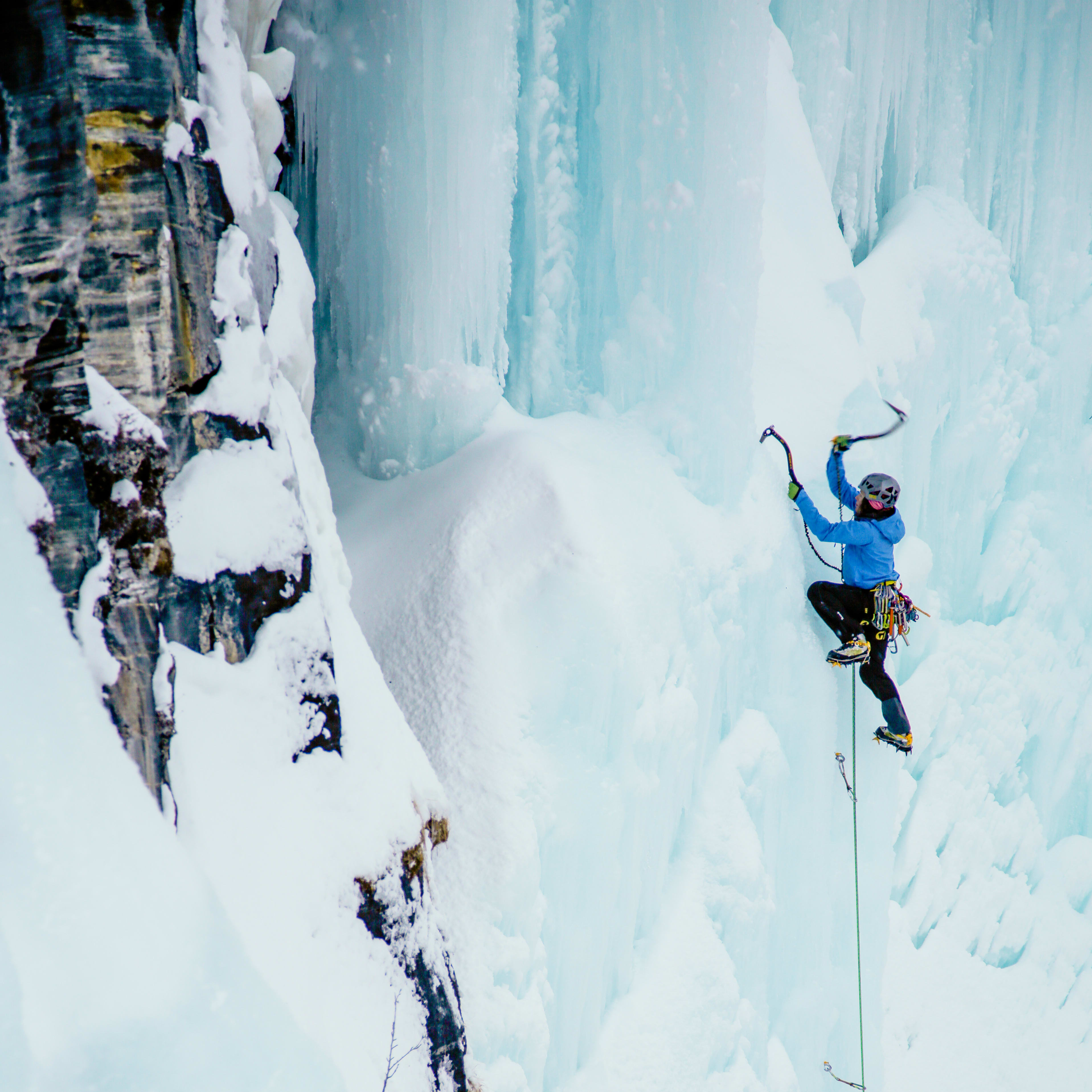 A climber wearing a blue Norrøna jacket ascends an icy cliff.  The climber is attached to a rope, and their ice axe is secured to the cliff. 
