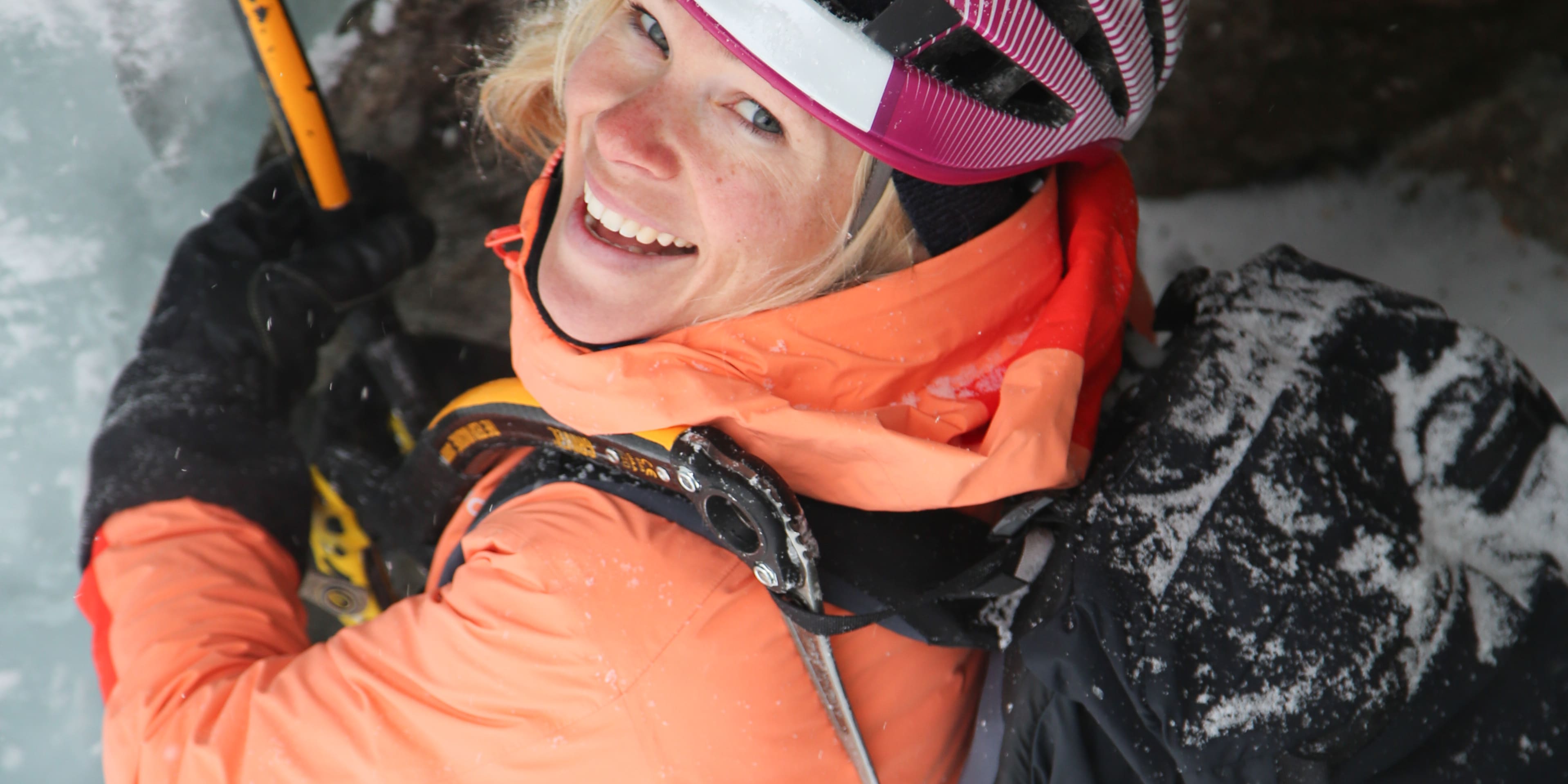 A smiling woman ice climbing, wearing a Norrøna jacket, helmet, and harness, with an ice ax and a rope. 
