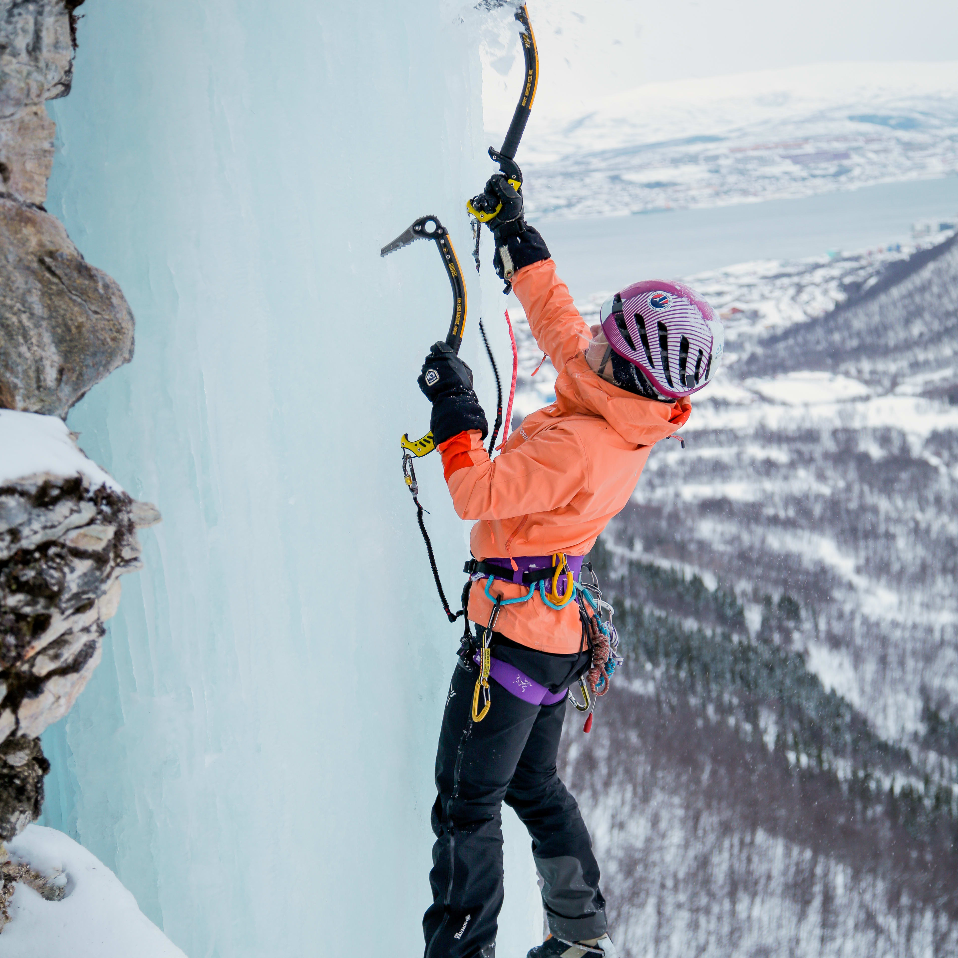 A climber wearing a Norrøna jacket and ice climbing gear ascends a snowy ice wall. 
