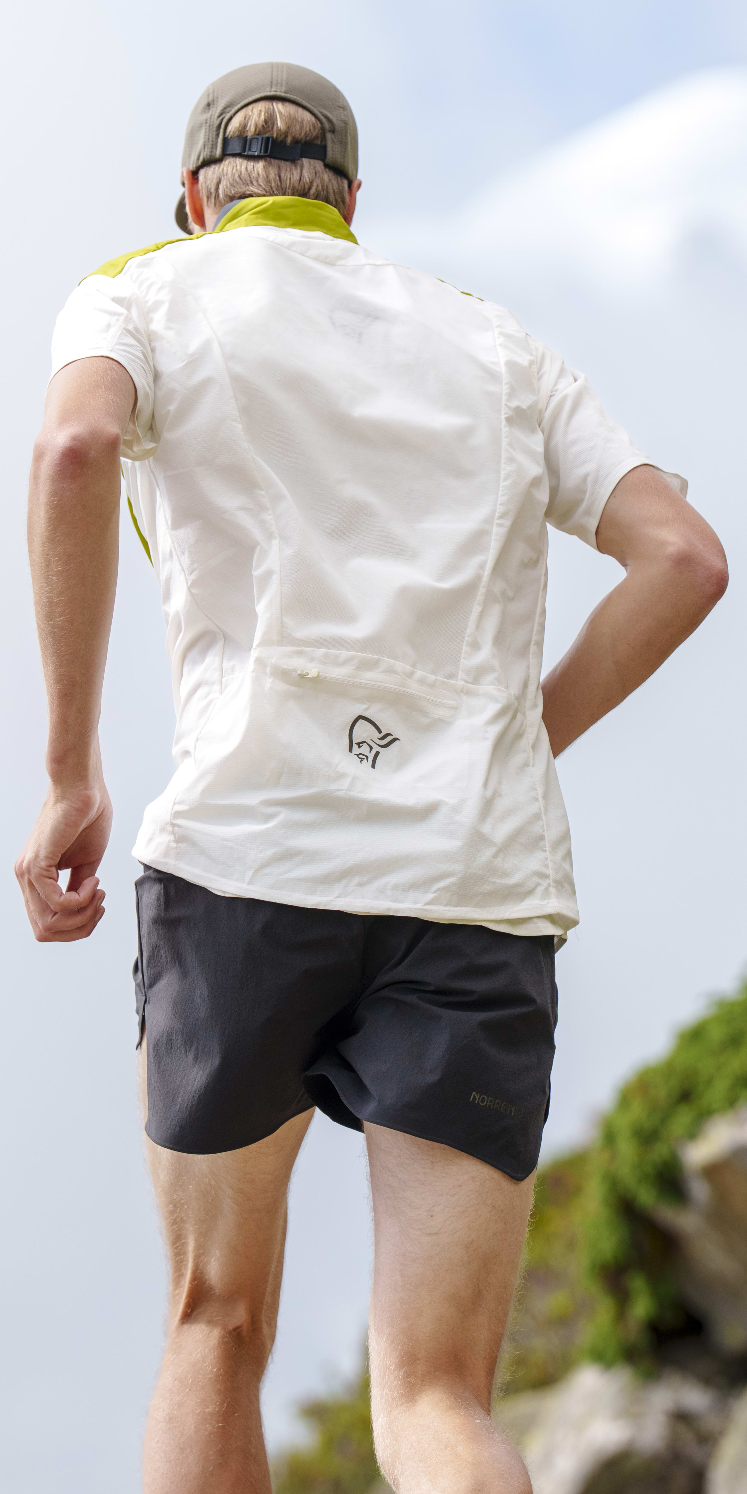 A man walks away, wearing a white Norrøna shirt, grey Norrøna shorts, and a hat.