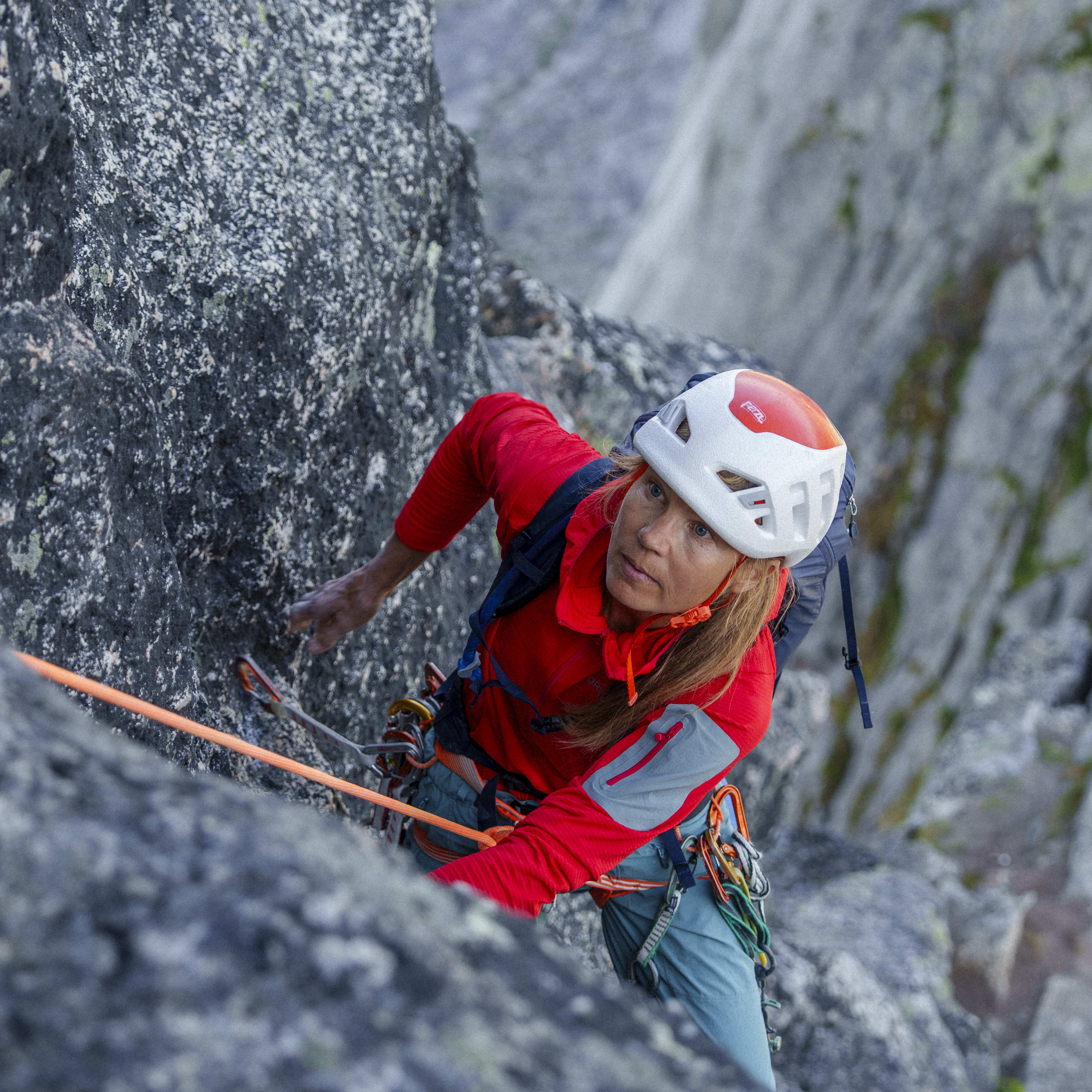 A climber in a red Norrøna jacket and white helmet ascends a rocky cliff, secured by rope, with a mountain backdrop.