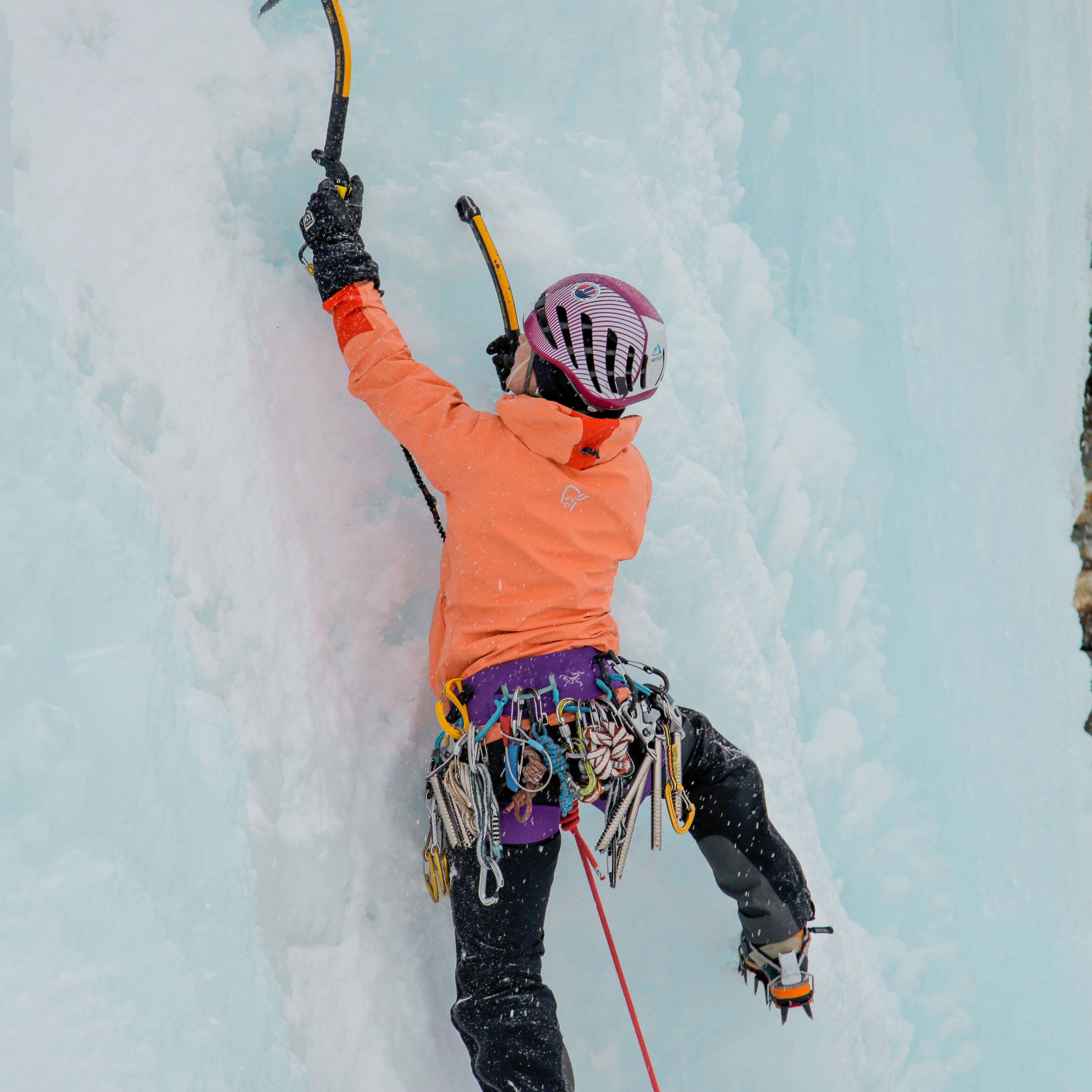A climber wearing a Norrøna jacket, helmet, and crampons ascends a wall of ice. 
