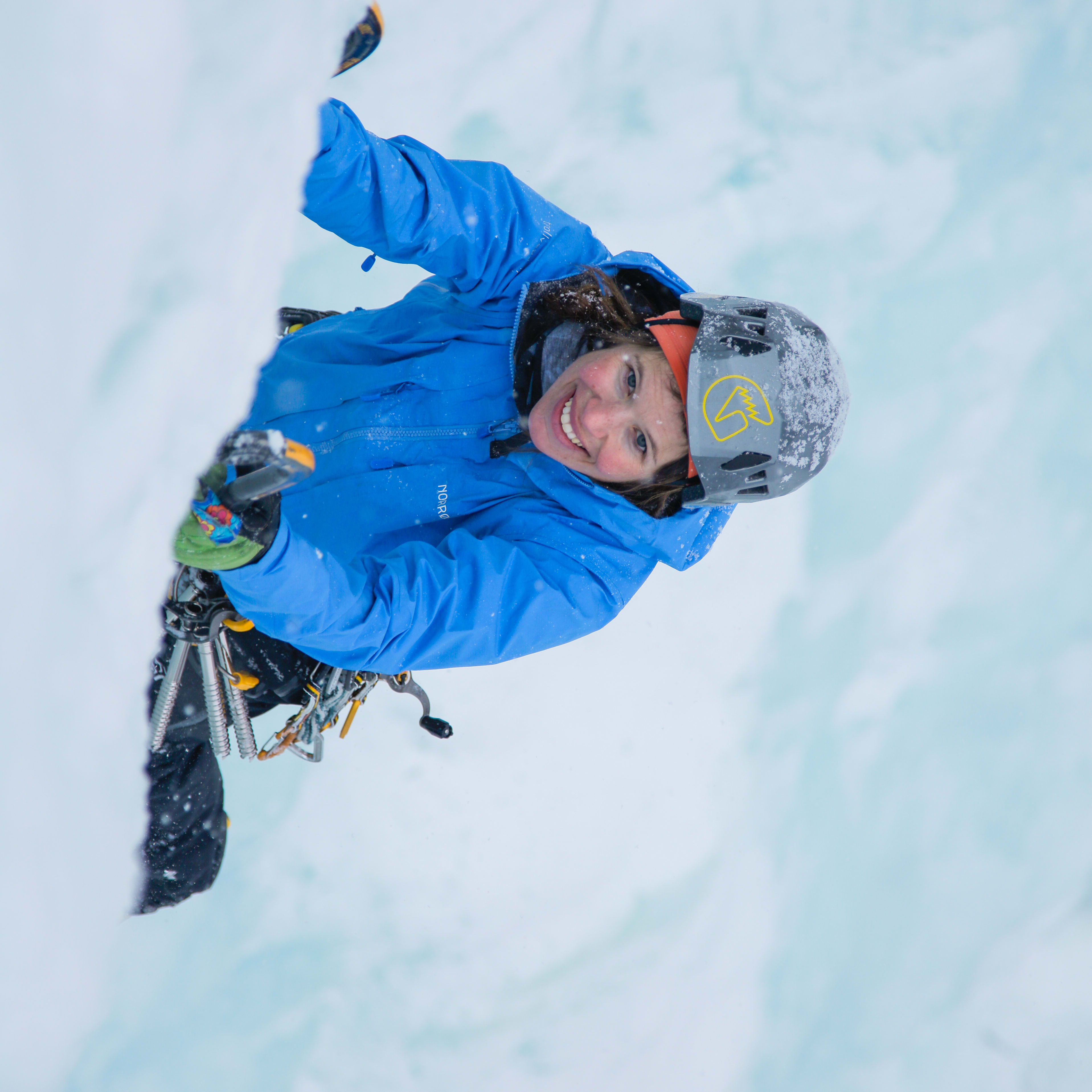 A woman wearing a blue Norrøna jacket and a helmet ice climbs a snowy wall. She is smiling at the camera.  
