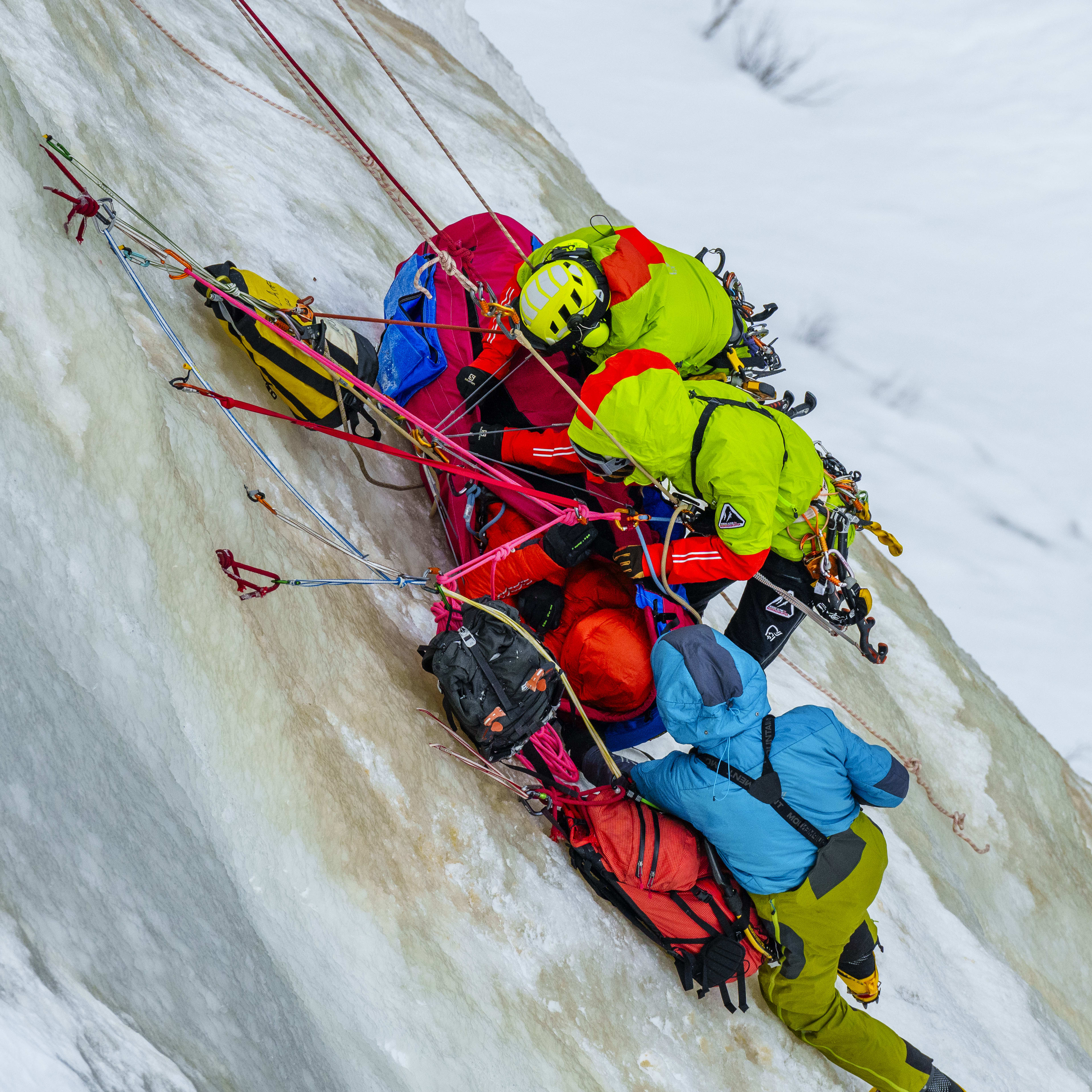 Two climbers are rappelling down an icy cliff. One is securing a person in a rescue harness, the other is belaying. Both are wearing Norrøna jackets and climbing gear.