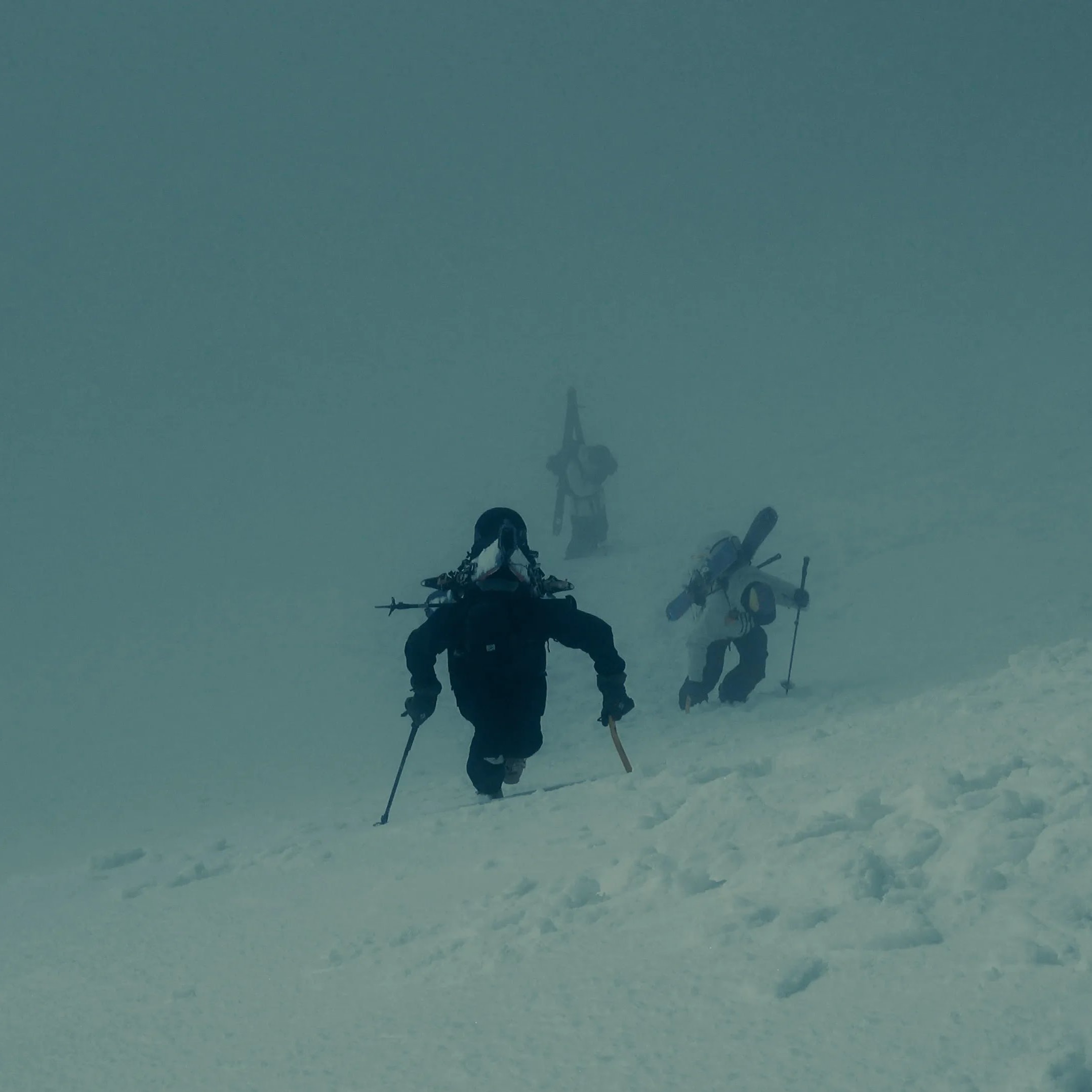 Three skiers in Norrøna gear climb a snowy slope in heavy fog.