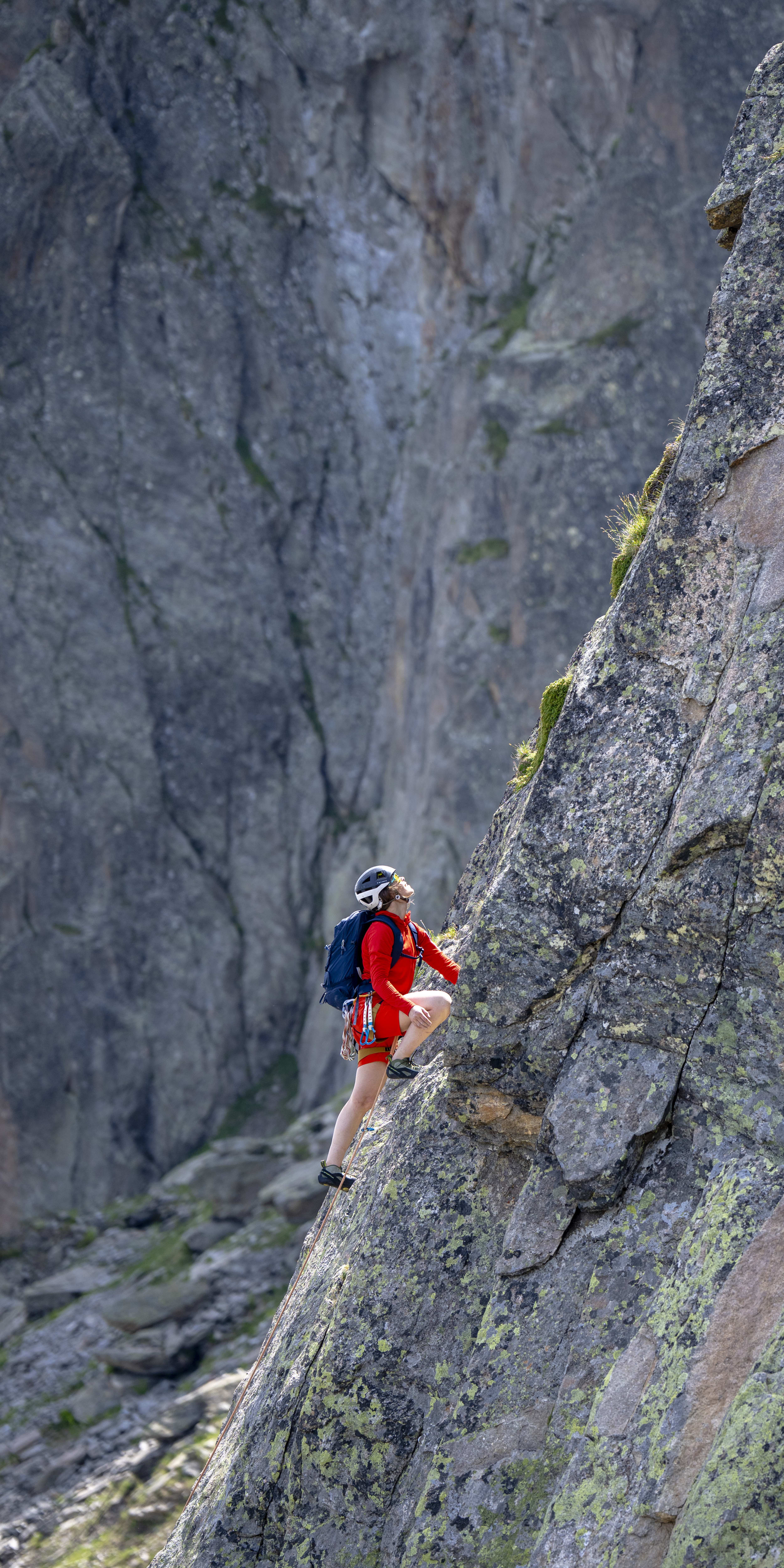 A rock climber in red Norrøna gear ascends a steep, lichen-covered rock face. The person wears a helmet and backpack with dramatic mountain backdrop.