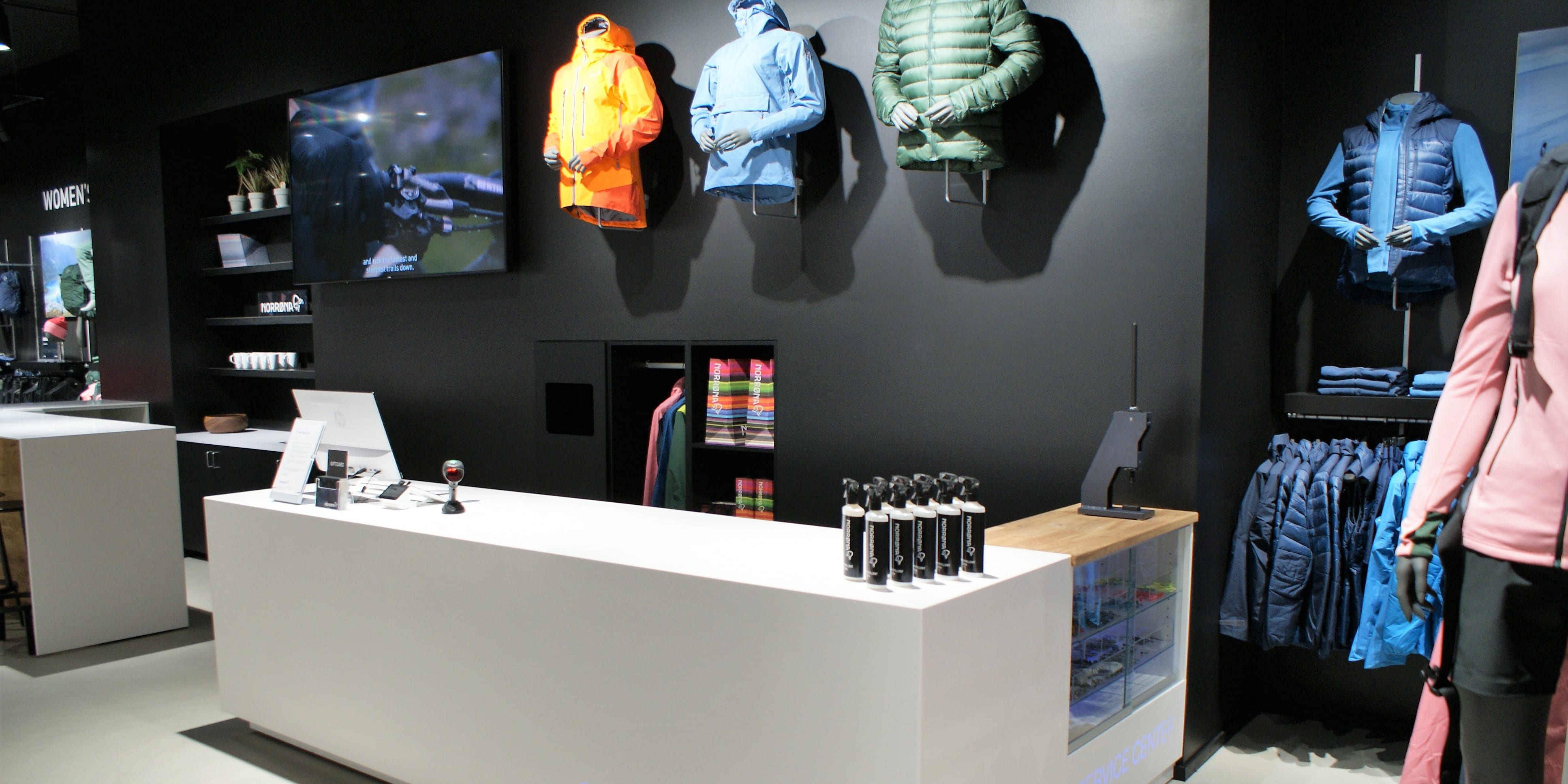 A white counter with the Norrøna logo in a retail store. The counter has bottles of Norrøna product.  There are jackets displayed on mannequins in the background. 
