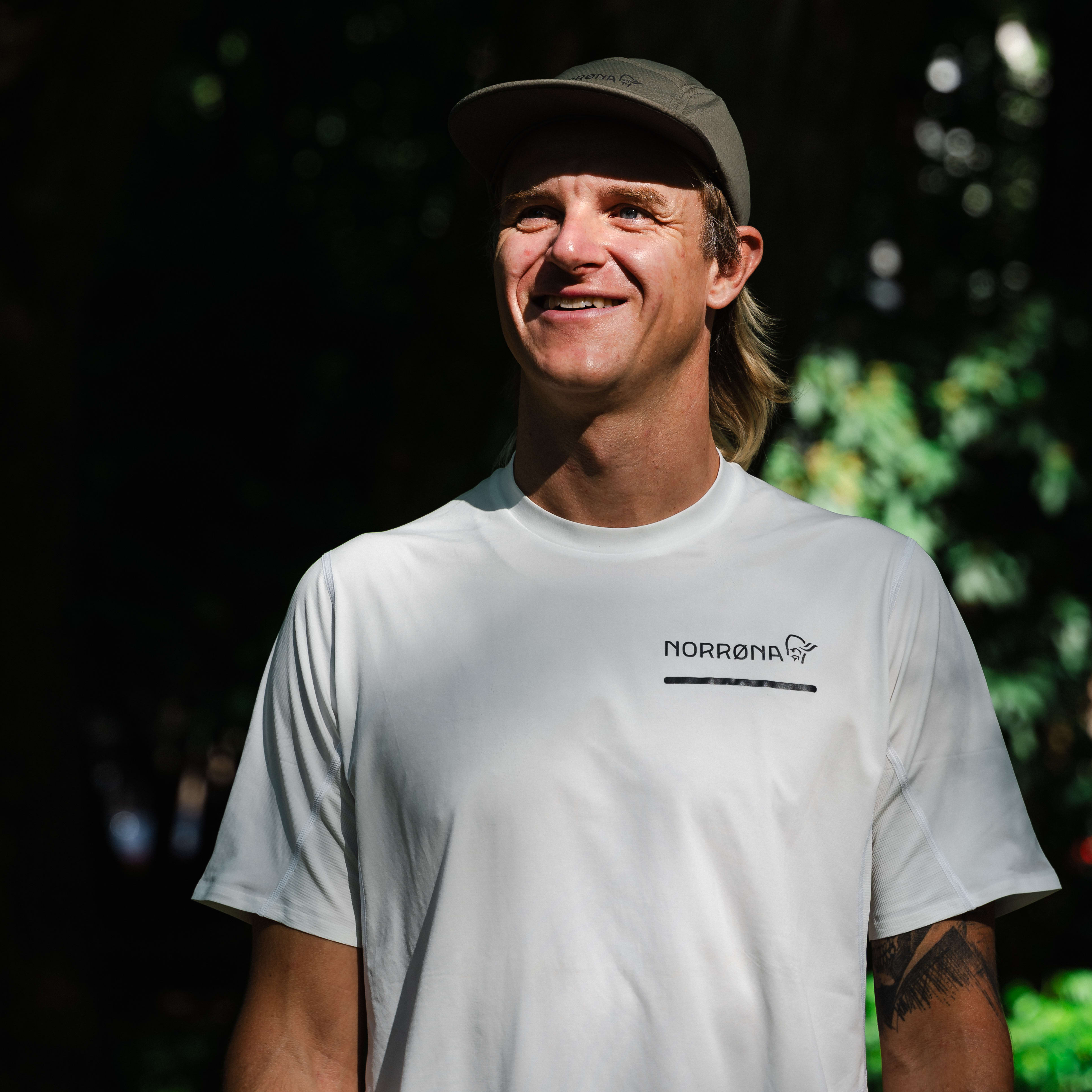 Smiling man in Norrøna tee and cap, outdoors in dappled sunlight.