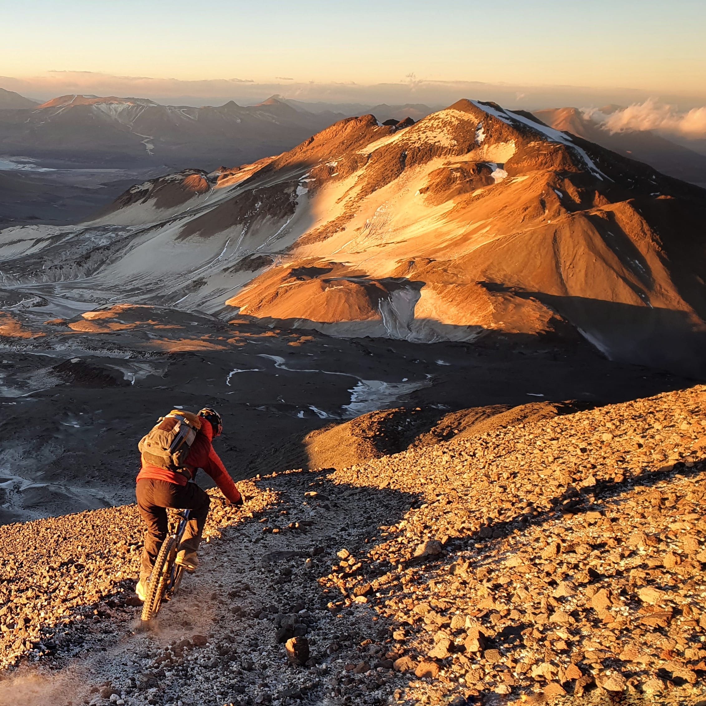 A person mountain biking down a rocky path in a mountainous landscape. The sun is setting behind the mountains, casting a warm glow on the scene. The biker is wearing a Norrøna jacket and riding a mountain bike. 
