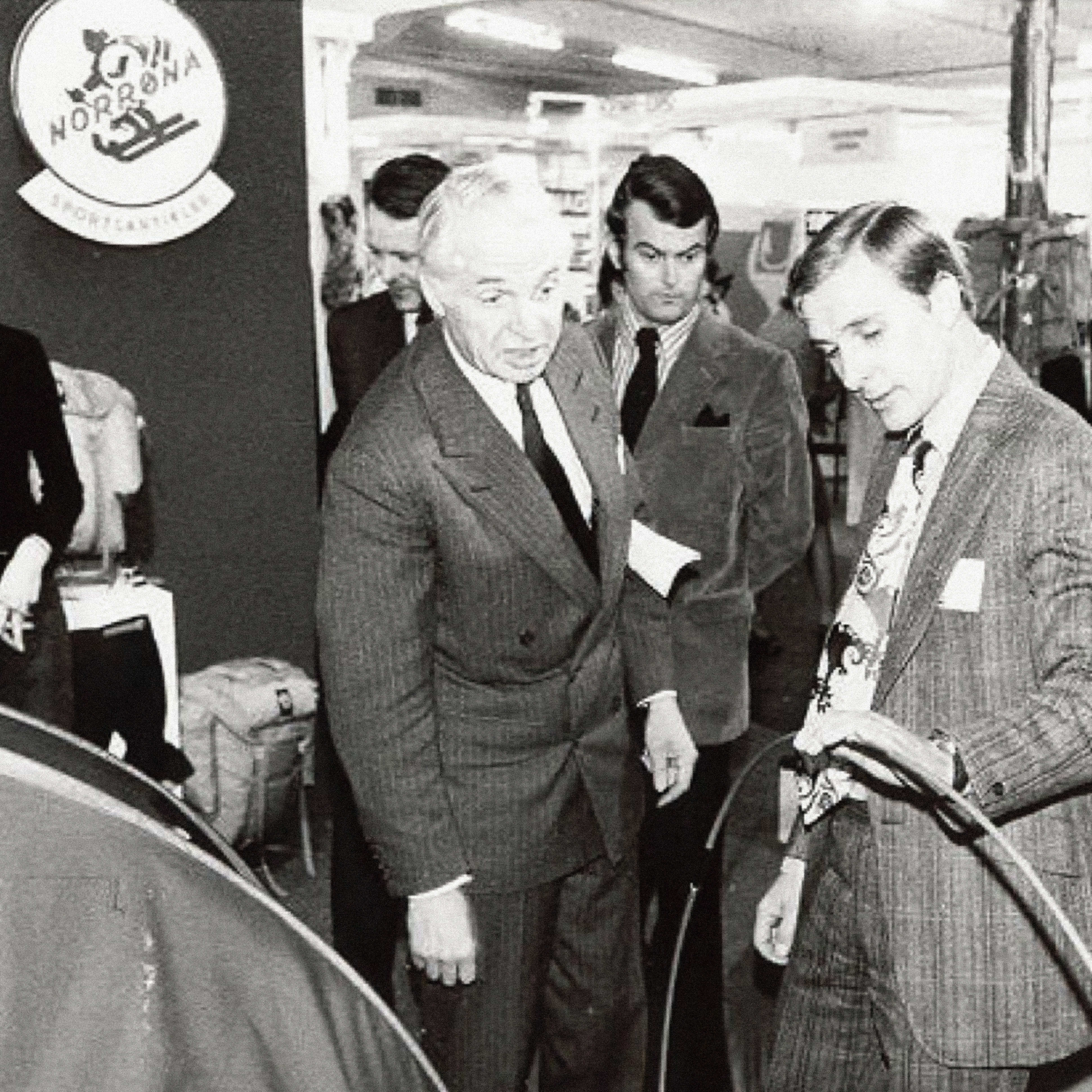 Black and white shot of Norrøna booth and its products. Men in suits inspect a tent. Backpacks sit on a table behind it. The men all wear suits.