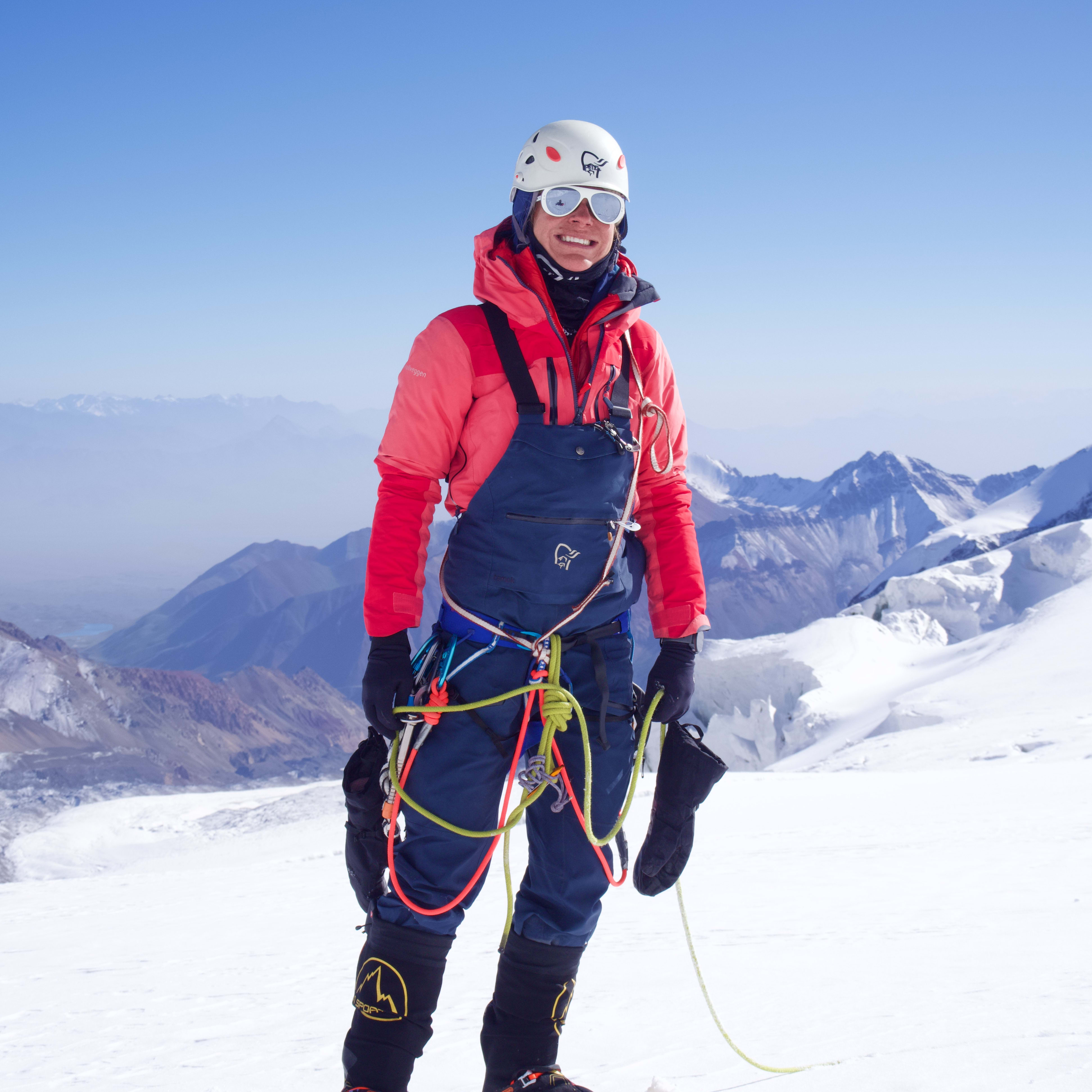 A smiling climber stands on snow against a mountain range, wearing a helmet, sunglasses, Norrøna shell jacket and bib, and mountaineering gear.

