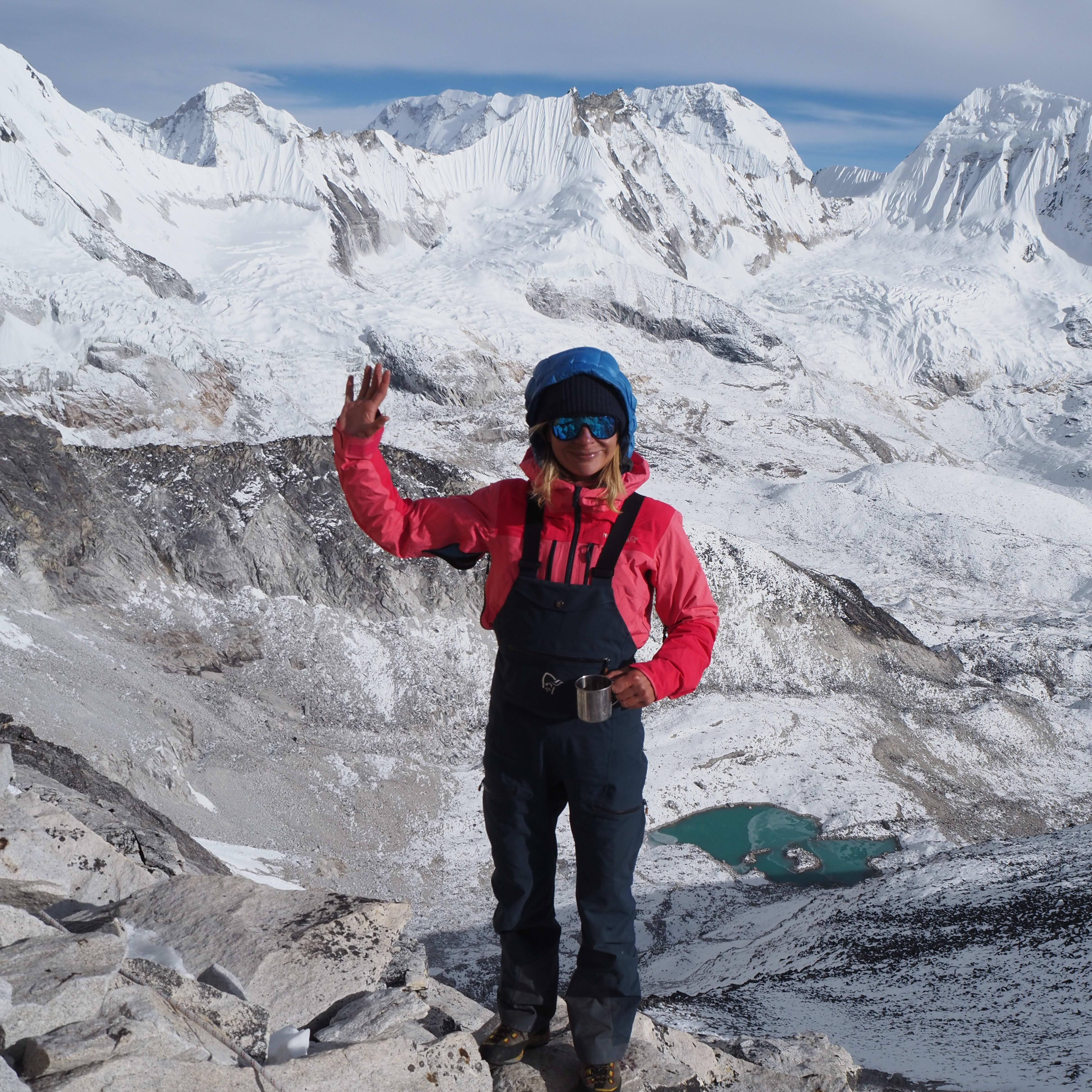 A woman wearing a Norrøna jacket and pants waves in the Himalayas. She stands on a snowy mountain with a turquoise lake in the background. 
