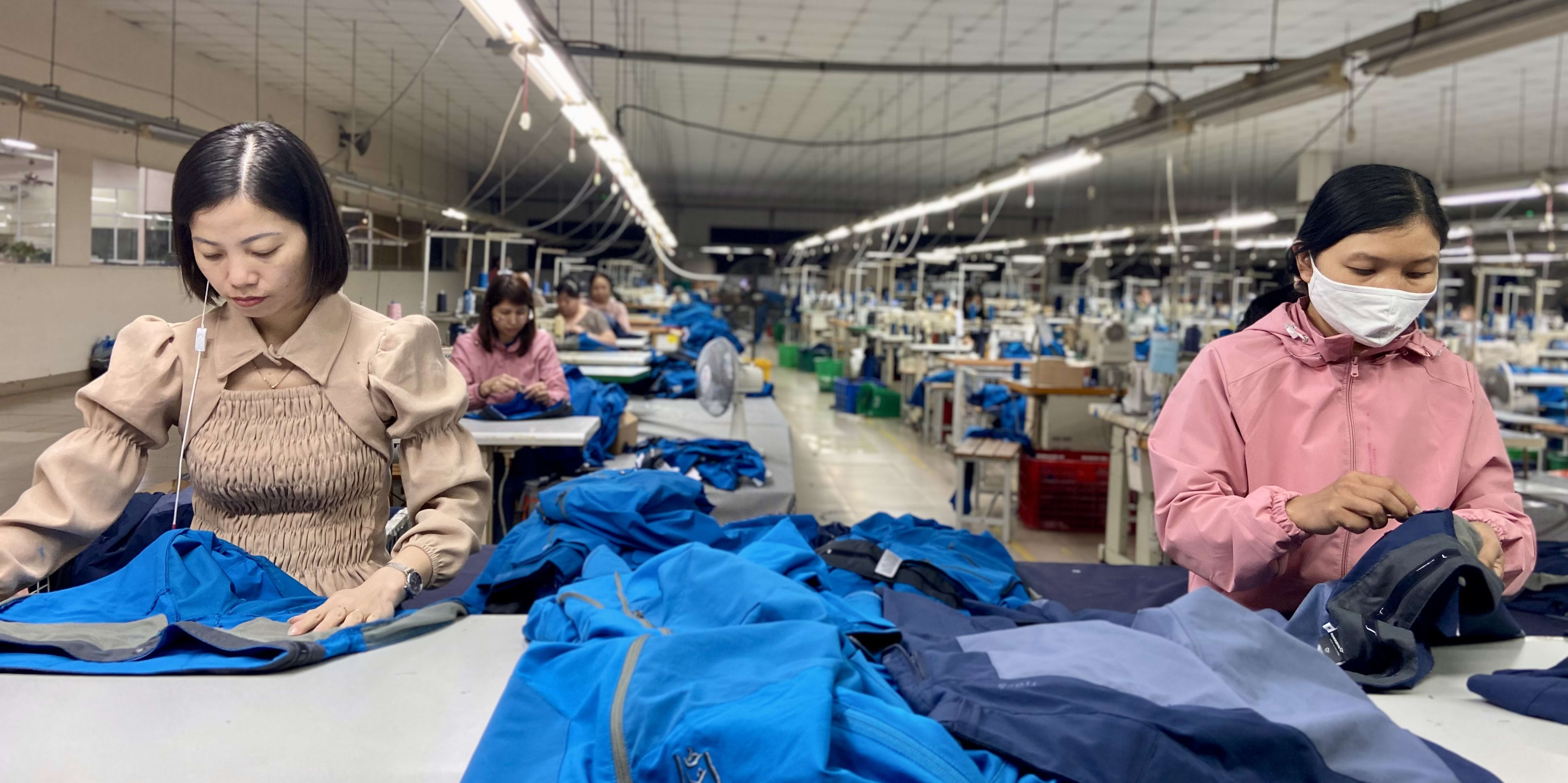 Two women working in a factory sewing Norrøna clothing.  A stack of blue jackets with the Norrøna logo is in the foreground. 
