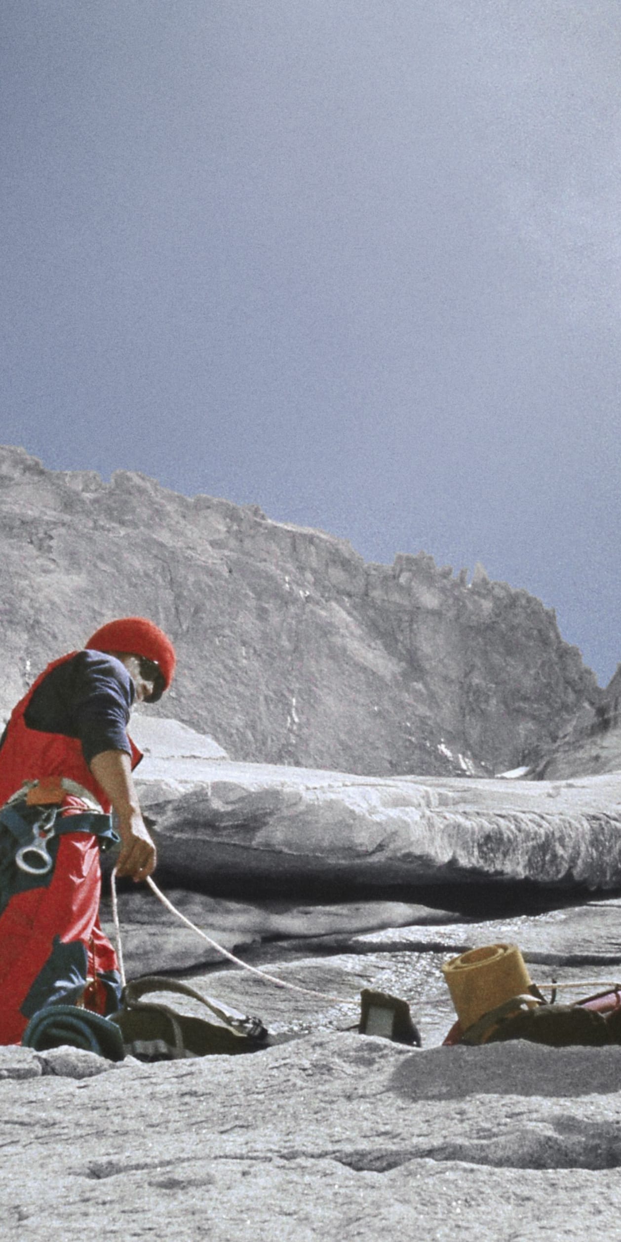 Three climbers, wearing red helmets and harnesses, ascend a rocky mountain slope.  The climbers are roped together, with gear visible.  A partially obscured fourth climber is seen below the other three.