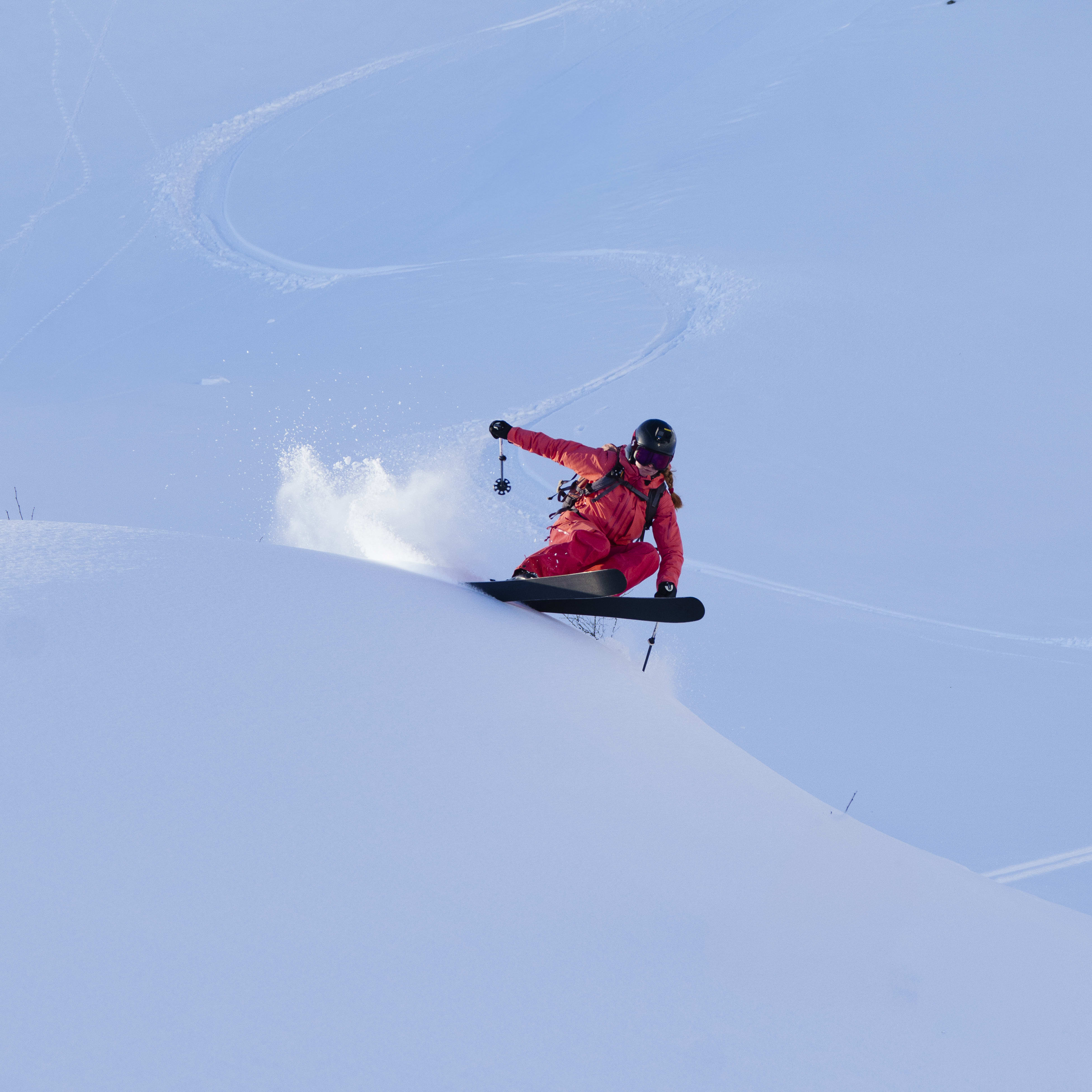 A snowboarder in Norrøna red outerwear jumps a snow-covered hill, kicking up powder against a blue sky.