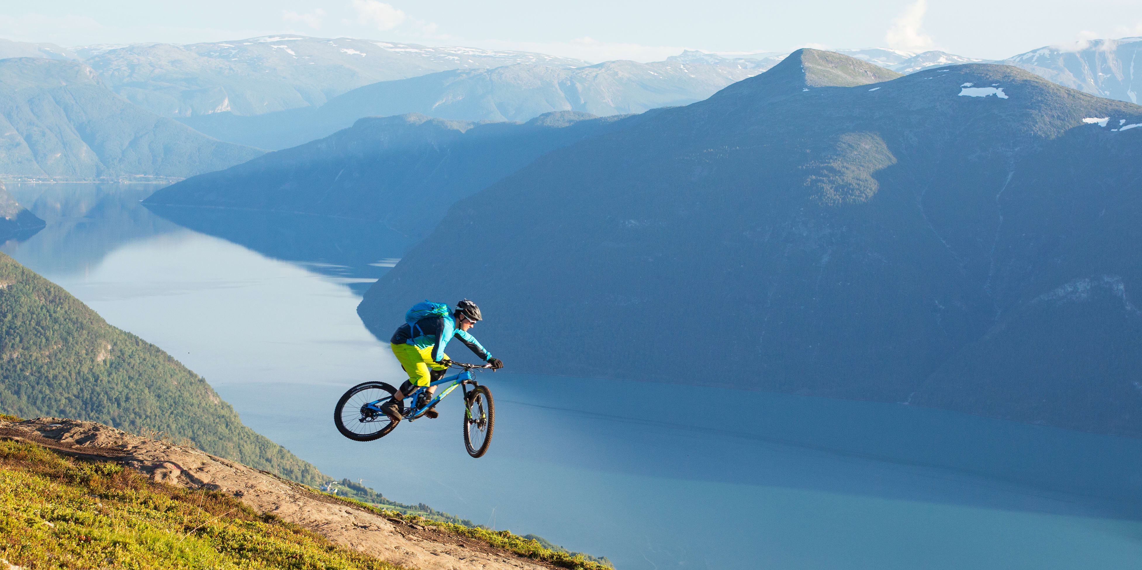 A person riding a Norrøna mountain bike off a cliff, overlooking a fjord and mountains.