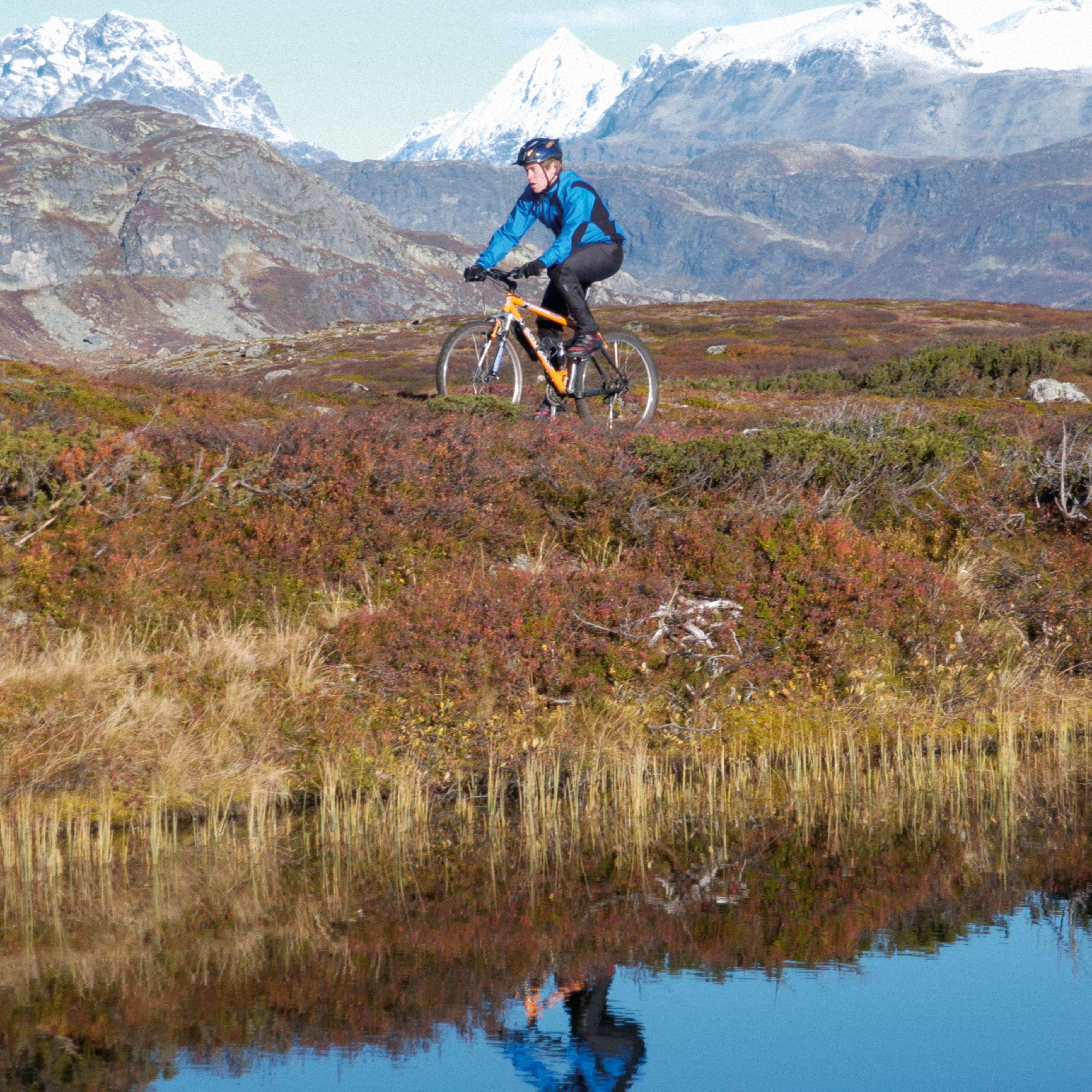 A person in a Norrøna jacket mountain biking in the highlands, with snow-capped mountains in the background, reflected in the water.