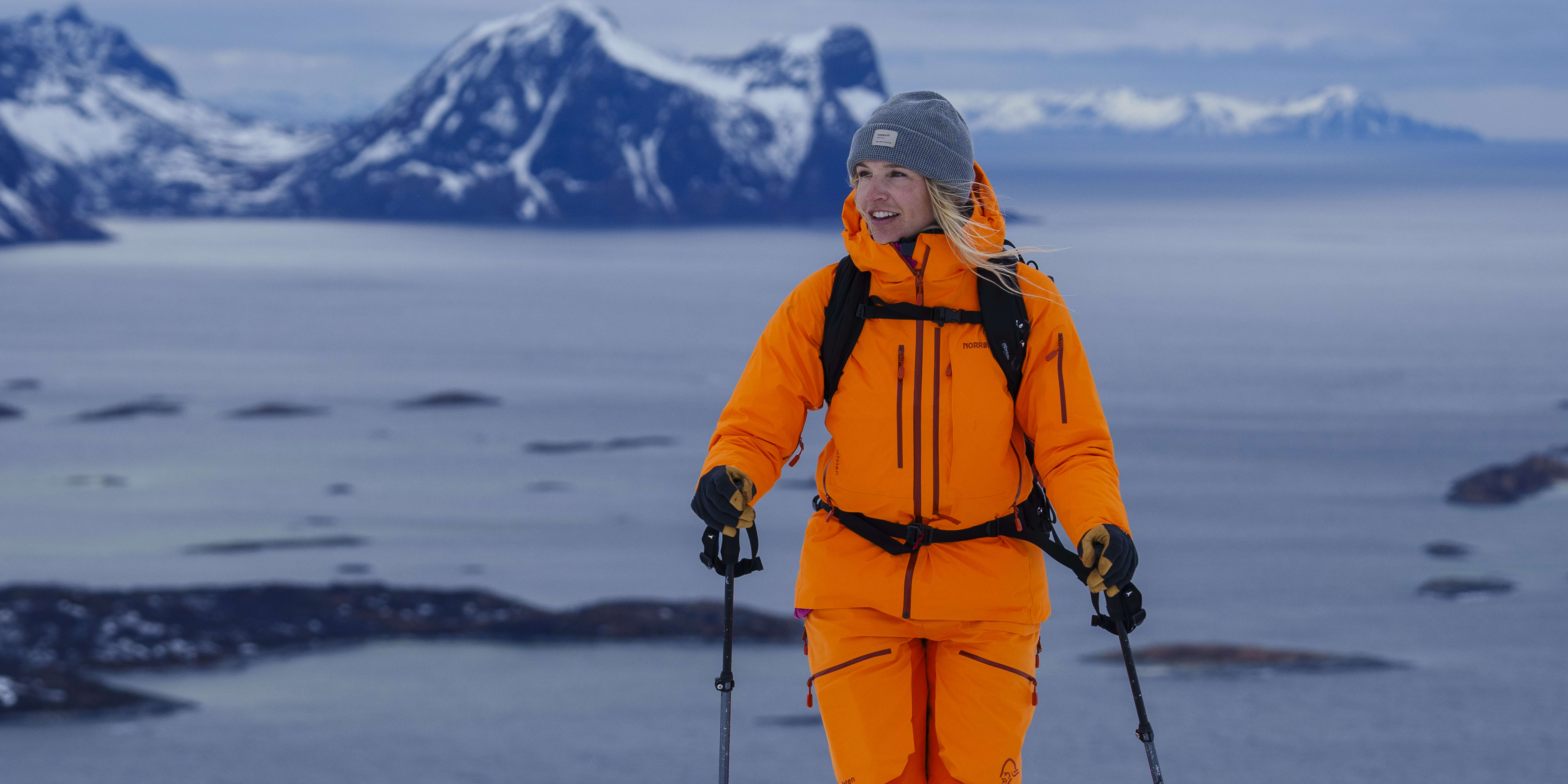 A woman wearing an orange Norrøna ski suit and a gray hat is standing on a snowy mountain with ski poles. In the background, you can see mountains and a body of water.