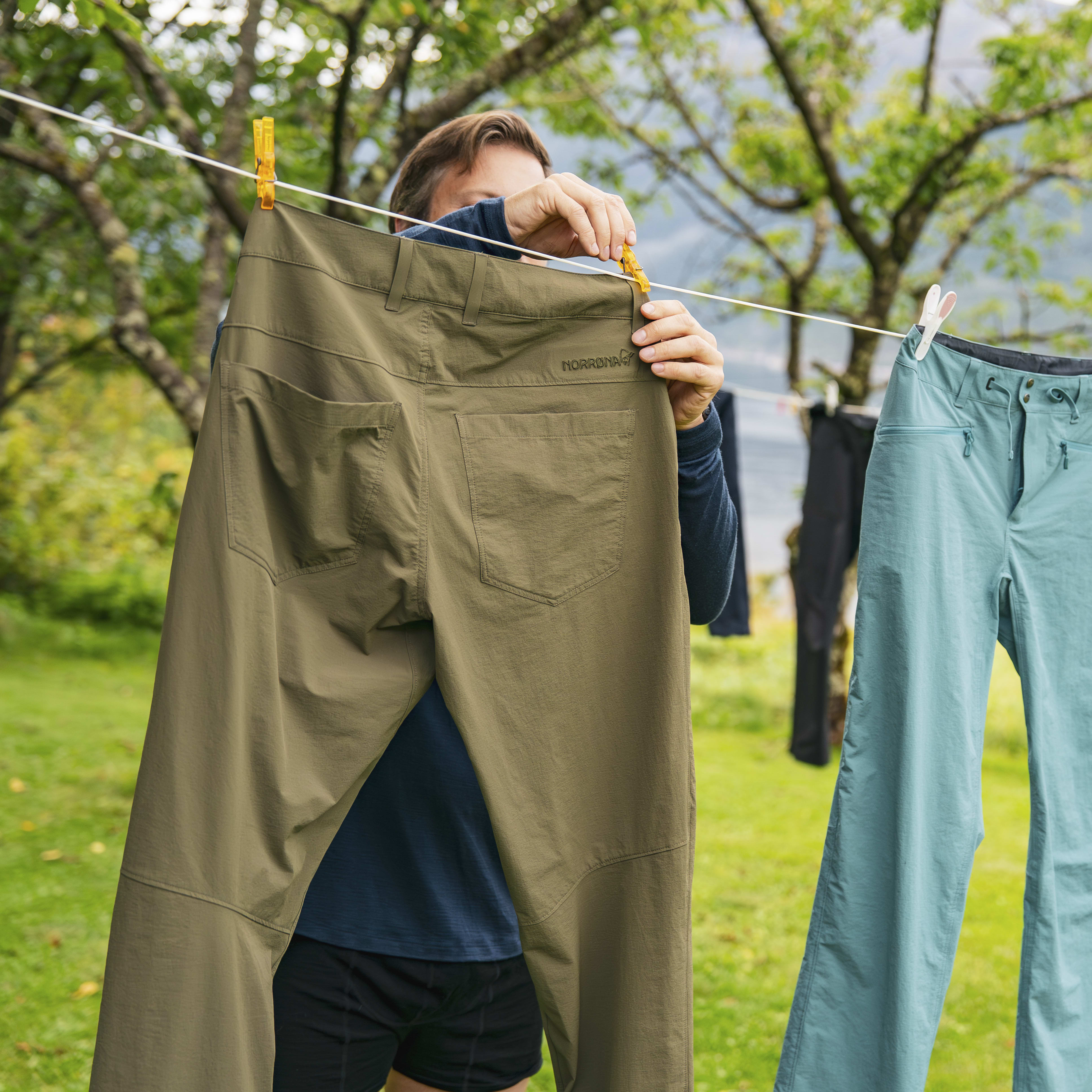 A person hangs Norrøna outdoor apparel on a clothesline with mountain views. Featured are khaki and blue pants, and a blue t-shirt.