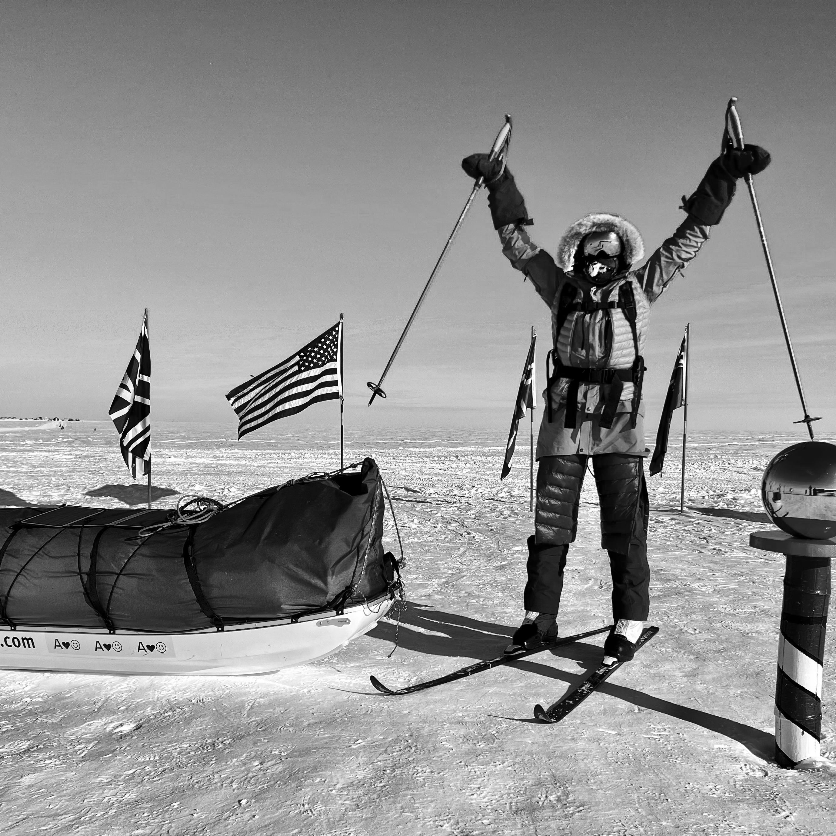 A person wearing Norrøna clothing stands on skis with arms raised, celebrating reaching the South Pole.