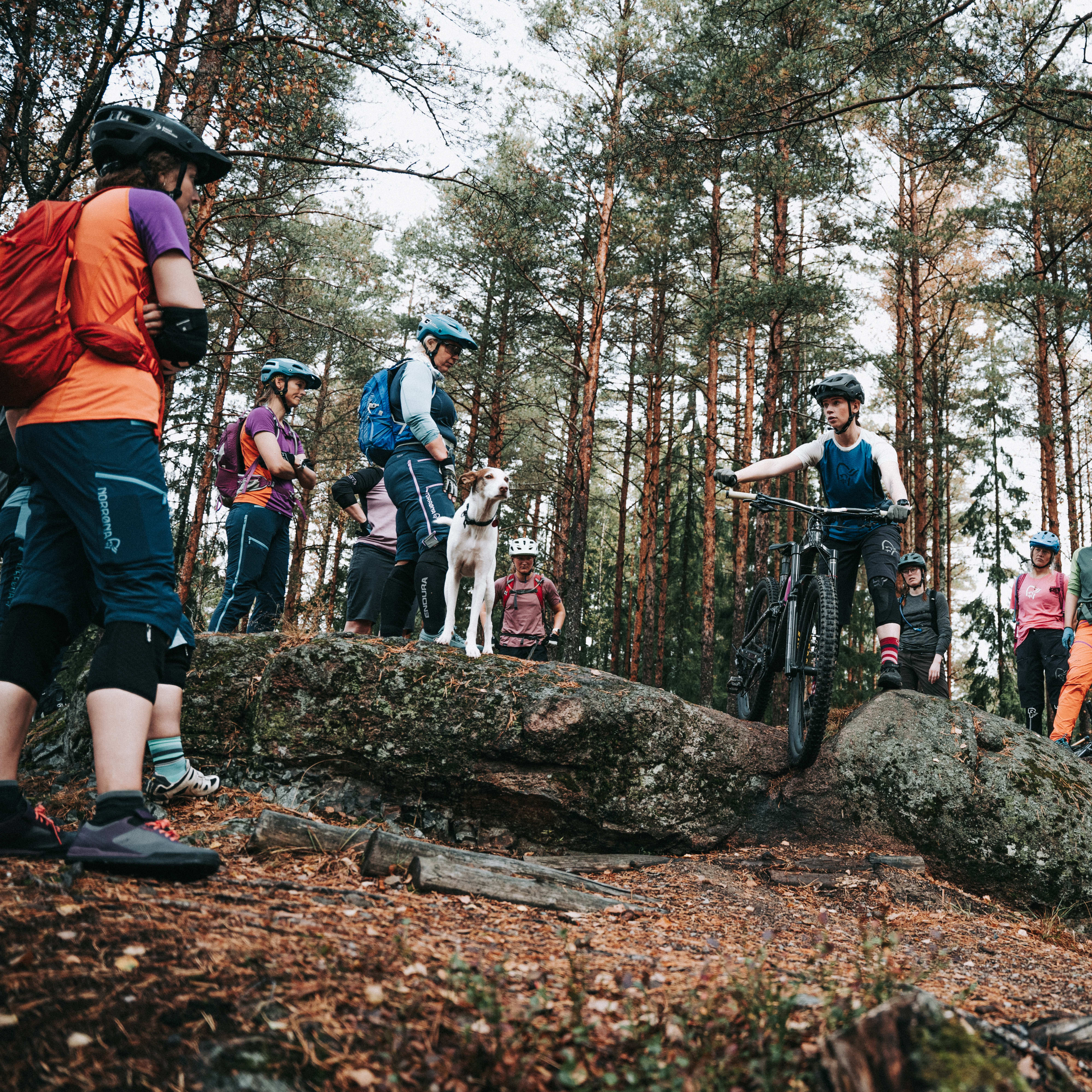 Group of people wearing helmets and Norrøna clothing in a forest setting with a mountain bike.