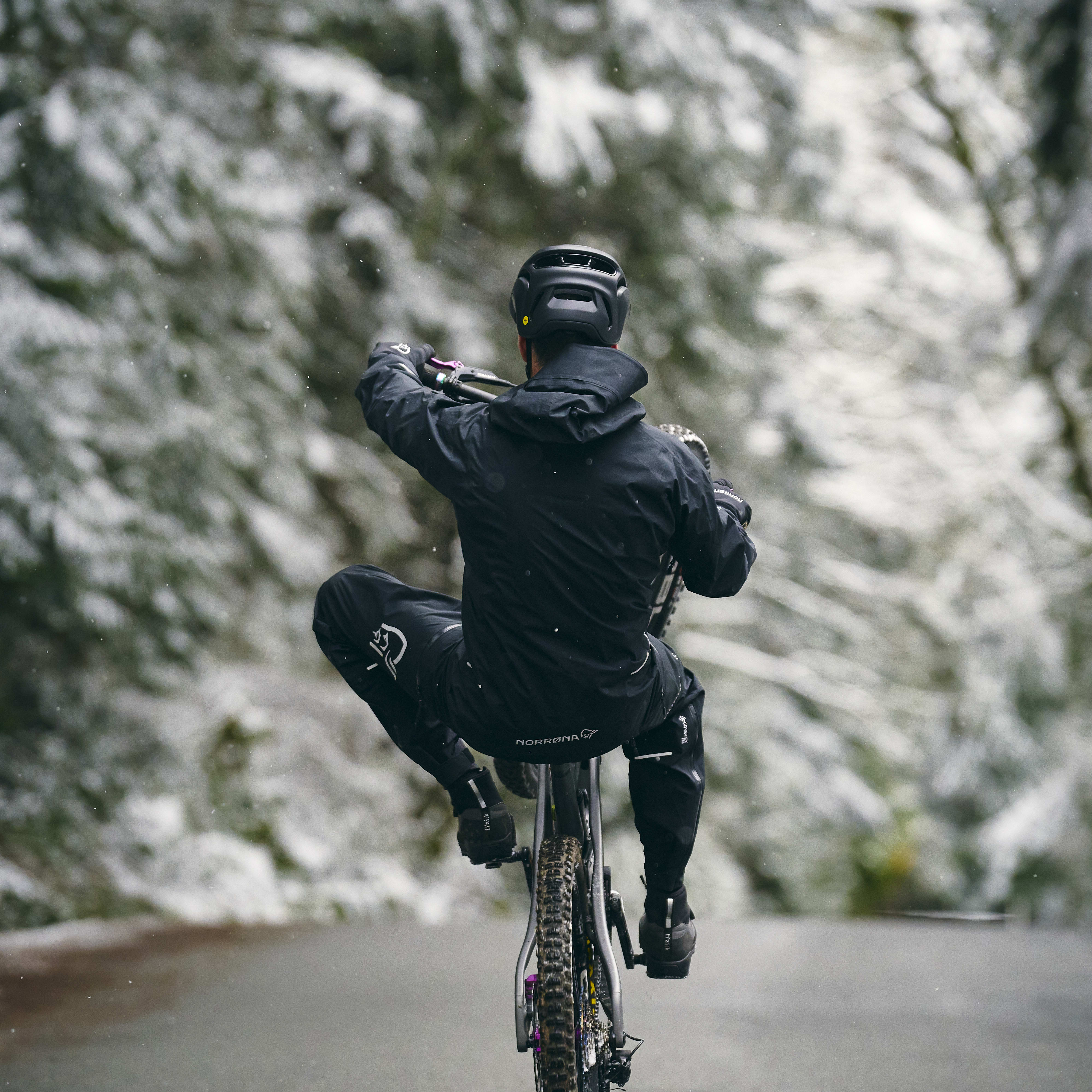 A person riding a bike on a snowy road, wearing a Norrøna jacket and pants.  The person is in the middle of doing a wheelie.