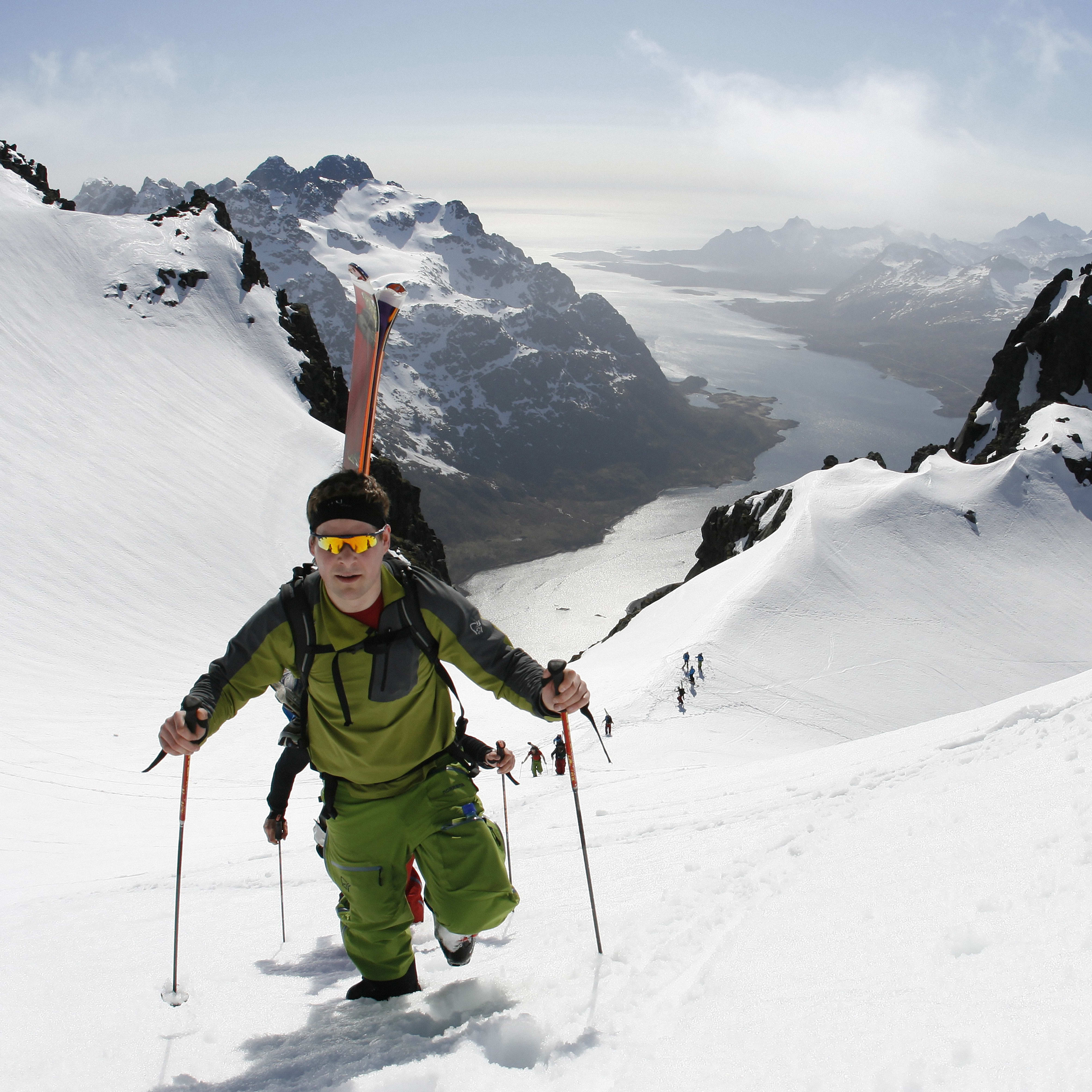 A man in Norrøna clothing skis up a snowy mountain with skis on his back, using poles for support. Mountains and water are in the background.