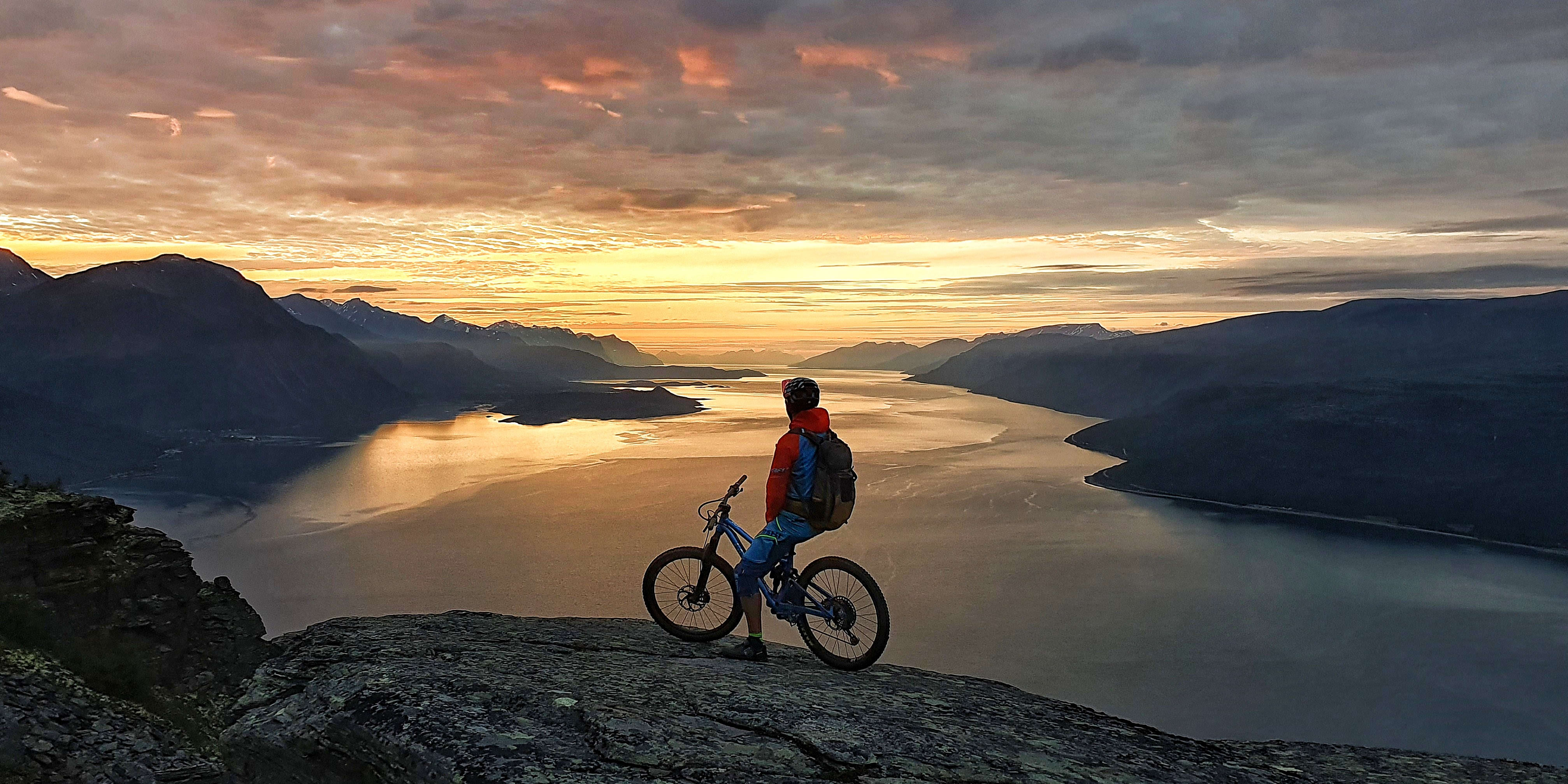 A person stands on a rocky cliff overlooking a large body of water. The person is facing away from the camera and is silhouetted against the golden sunrise. They are holding a mountain bike. The mountains can be seen in the distance.