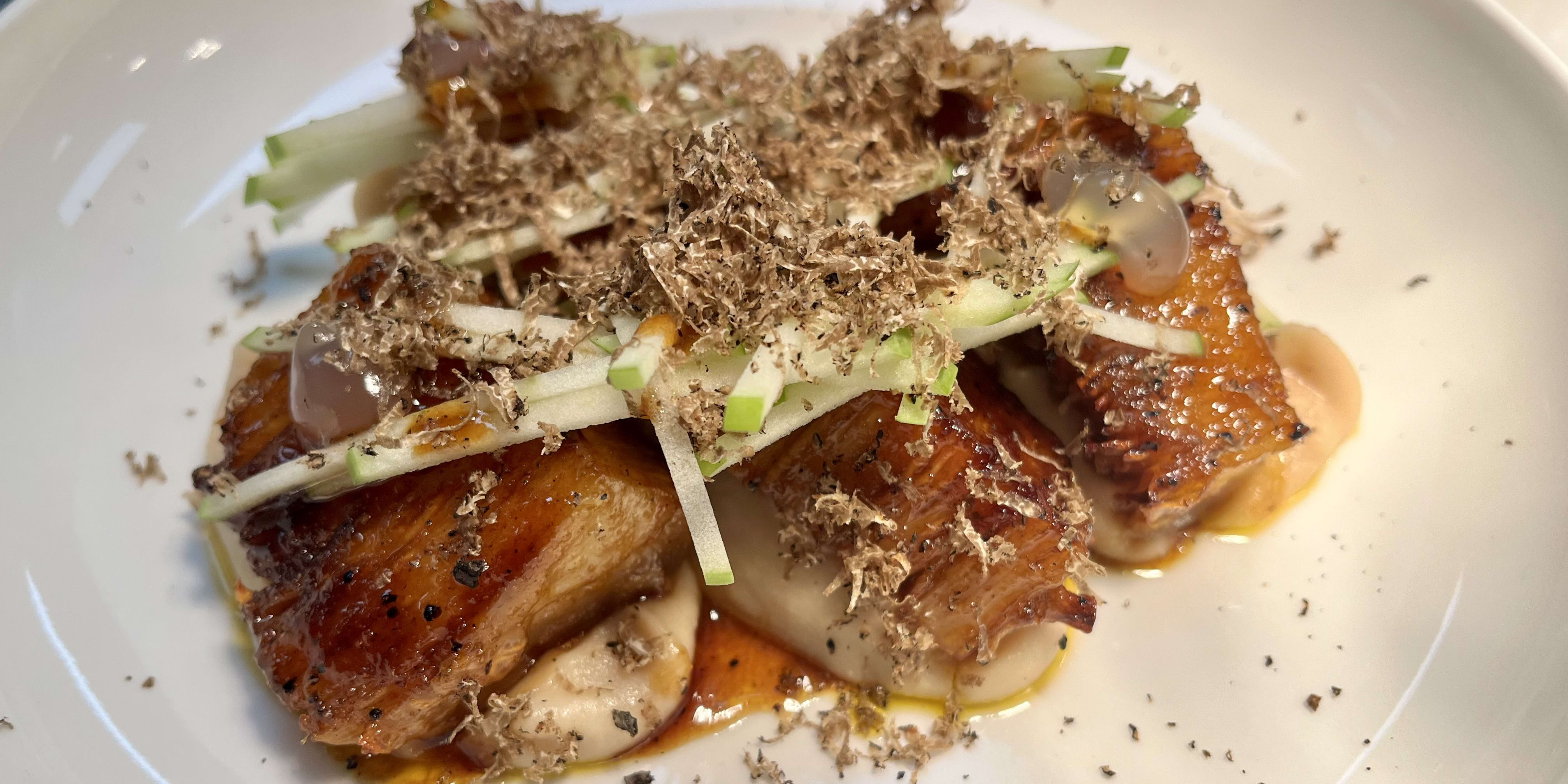 Close up of a plate of food. There are pieces of meat topped with shaved truffles, apple slices, and sauce.