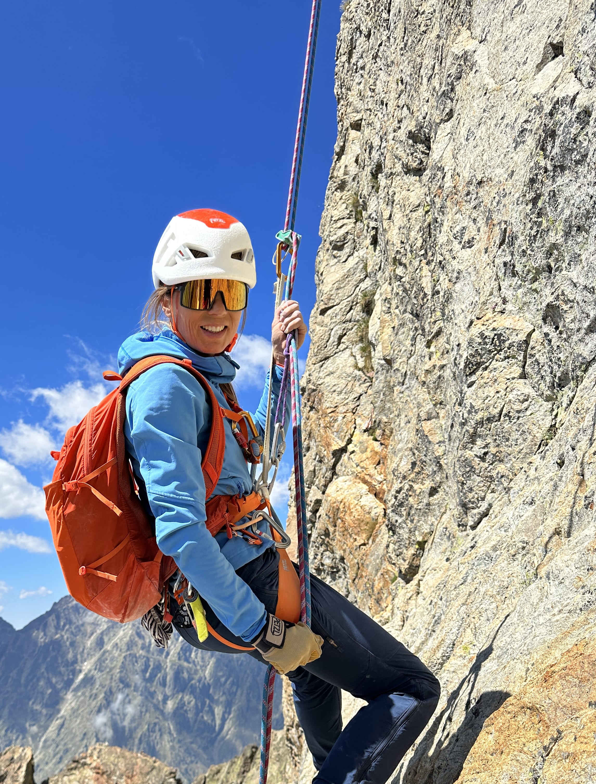 A woman wearing a helmet and Norrøna clothing rappels down a rocky cliff face. The background includes a blue sky and a mountain.
