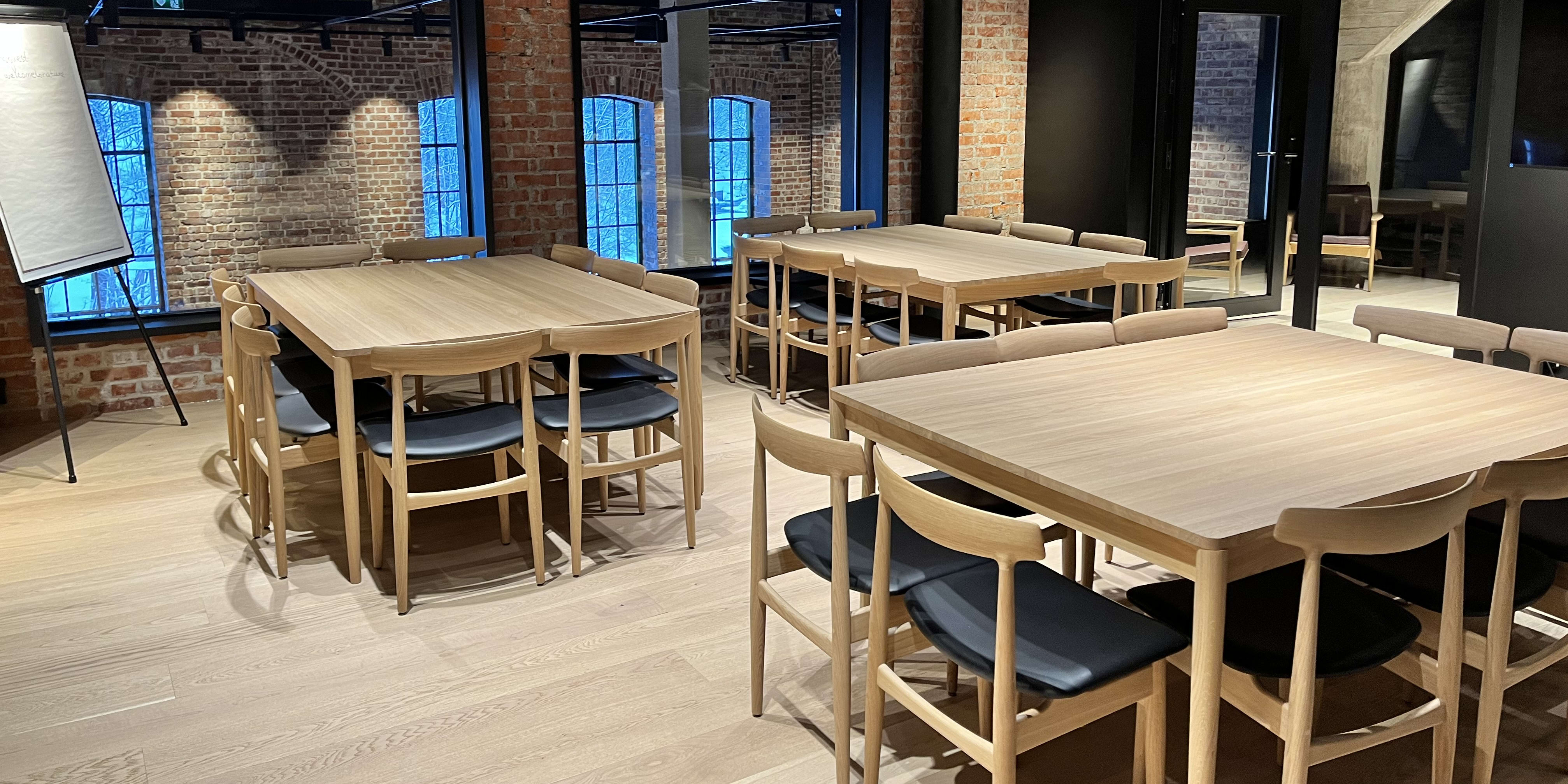 A meeting room with two large wooden tables and chairs. The chairs have black seats and are made by Norrøna. There is a whiteboard in the room and the windows show an outside view of a brick building. The room has a wood floor and a black tiled ceiling. 
