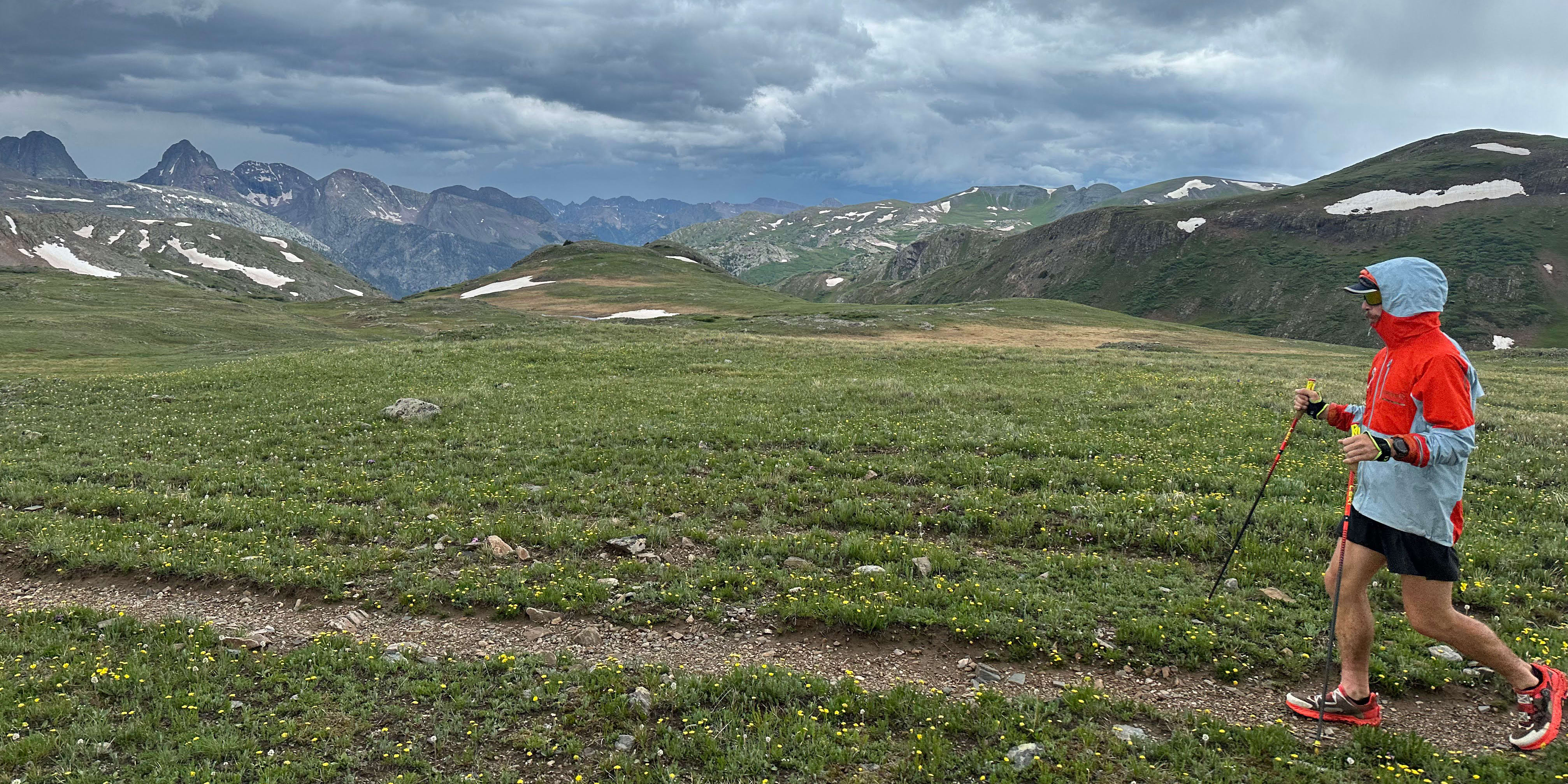 A hiker in a Norrøna jacket walks on a mountain path with a view of distant mountains. 
