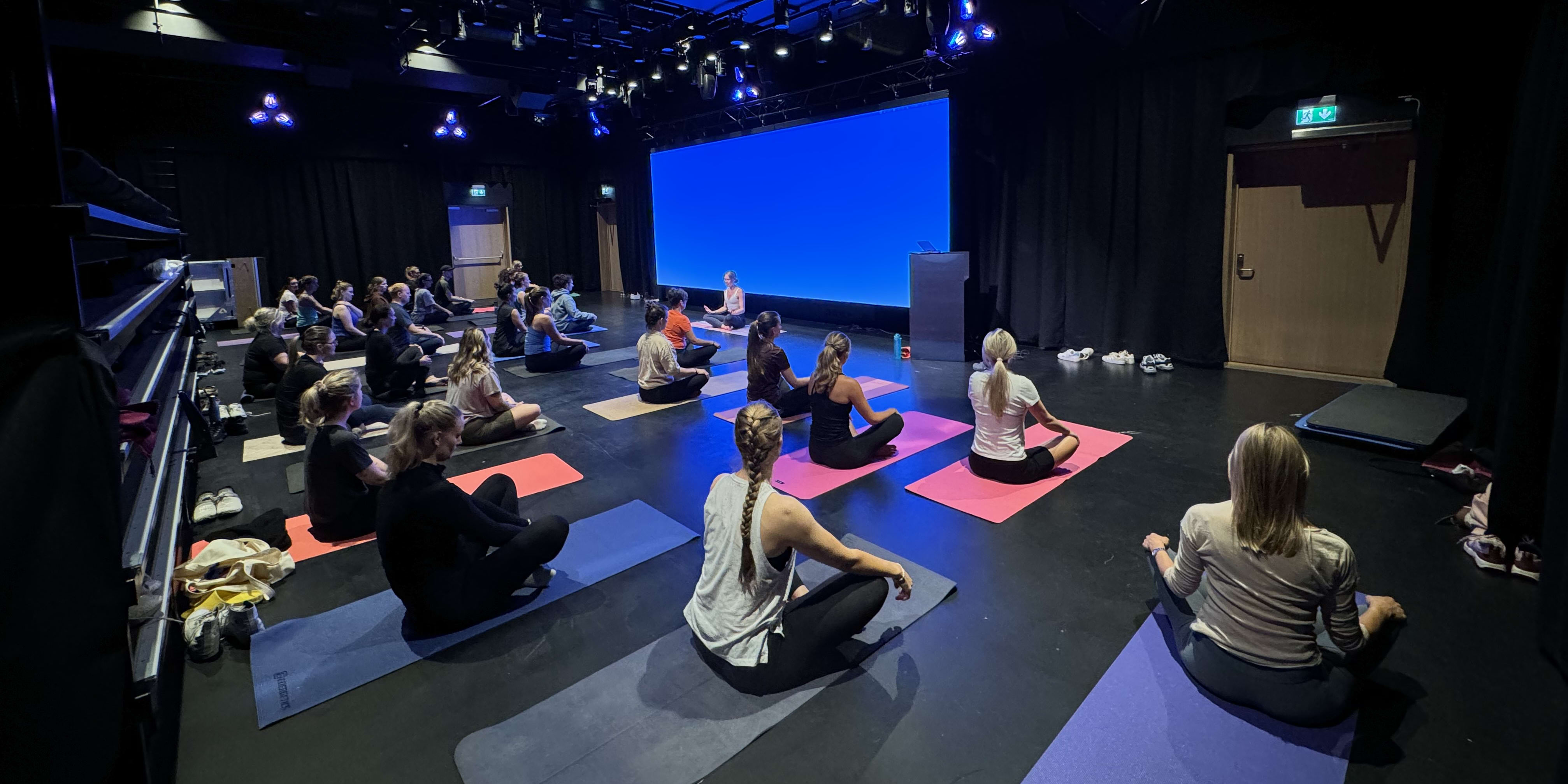 A group of people sitting in a yoga class. A large blue screen is behind them. Some are wearing Norrøna clothing.