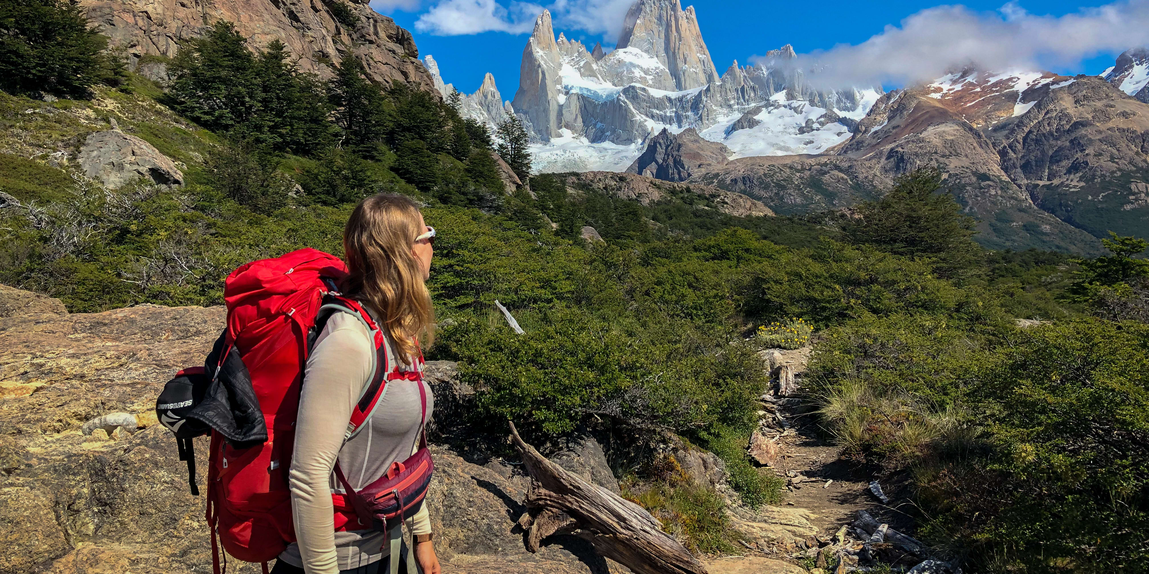 A woman wearing a Norrøna shirt and a red backpack looks up at the snow-capped mountains in the distance.