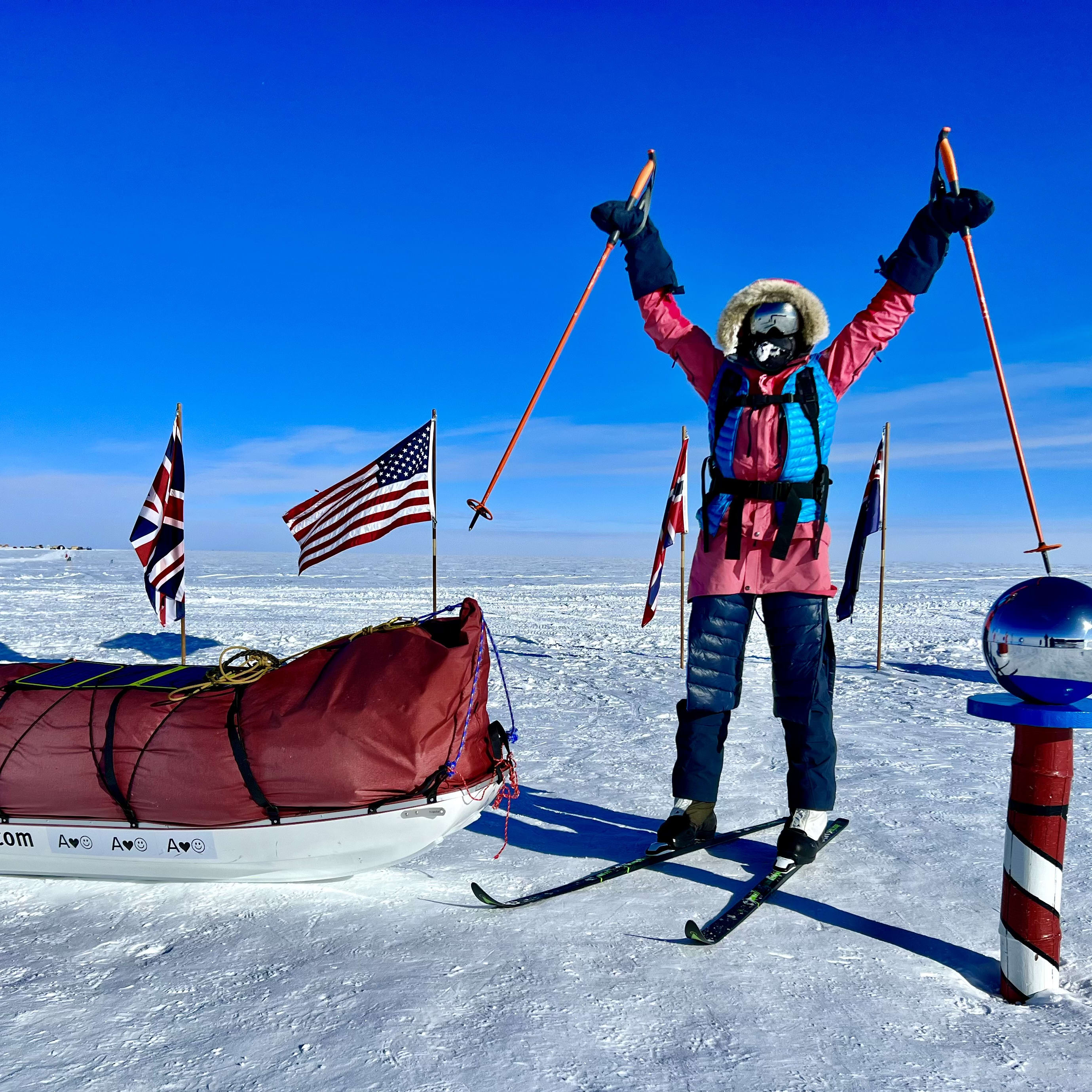 A person wearing Norrøna clothing is skiing with poles raised in the air against a blue sky. There are several flags and a sled in the background.  
