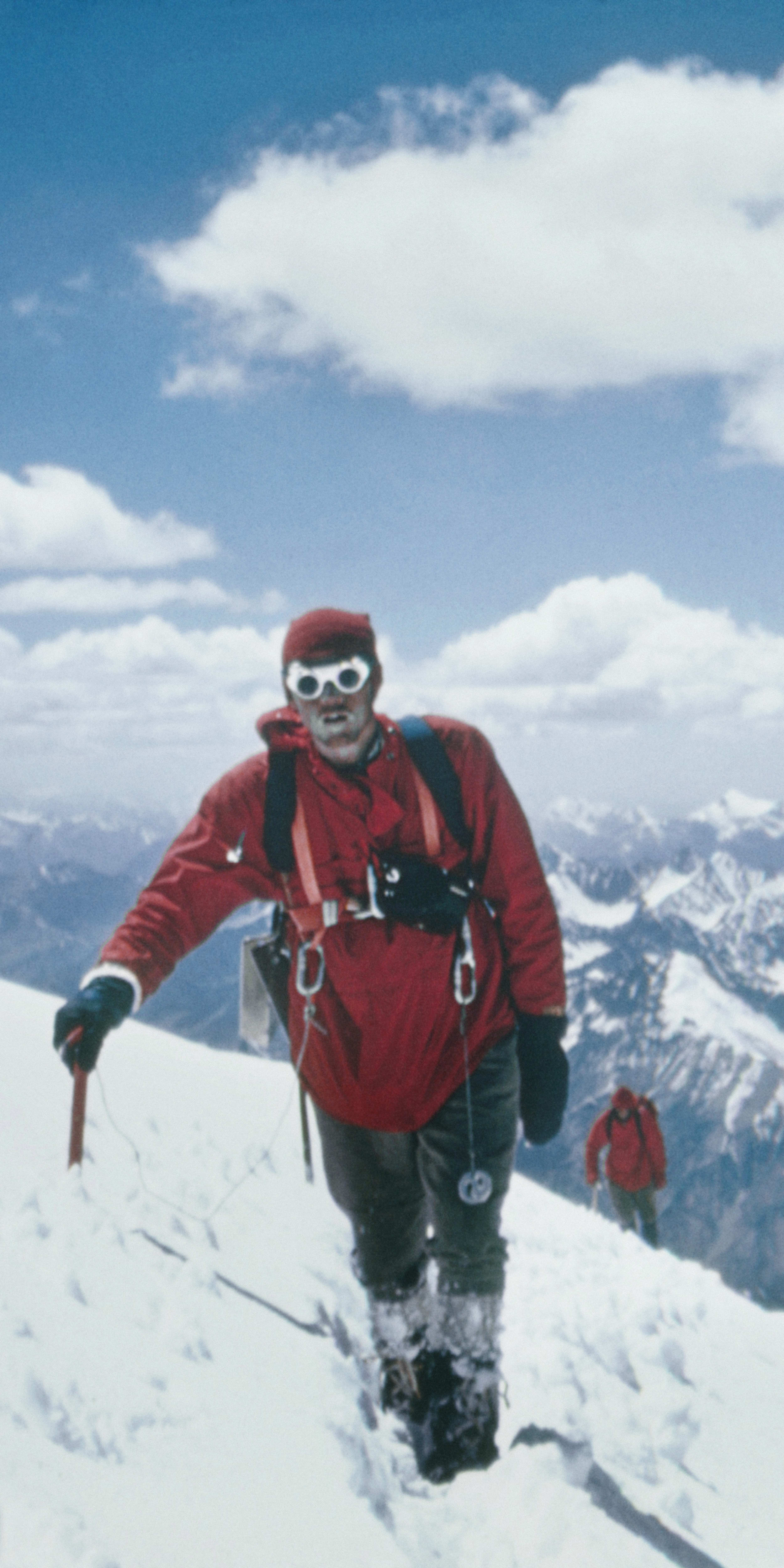 A man in red clothing, wearing goggles, is hiking across a snowy mountaintop. The mountains in the background are covered in snow. The sky is blue with white clouds.  