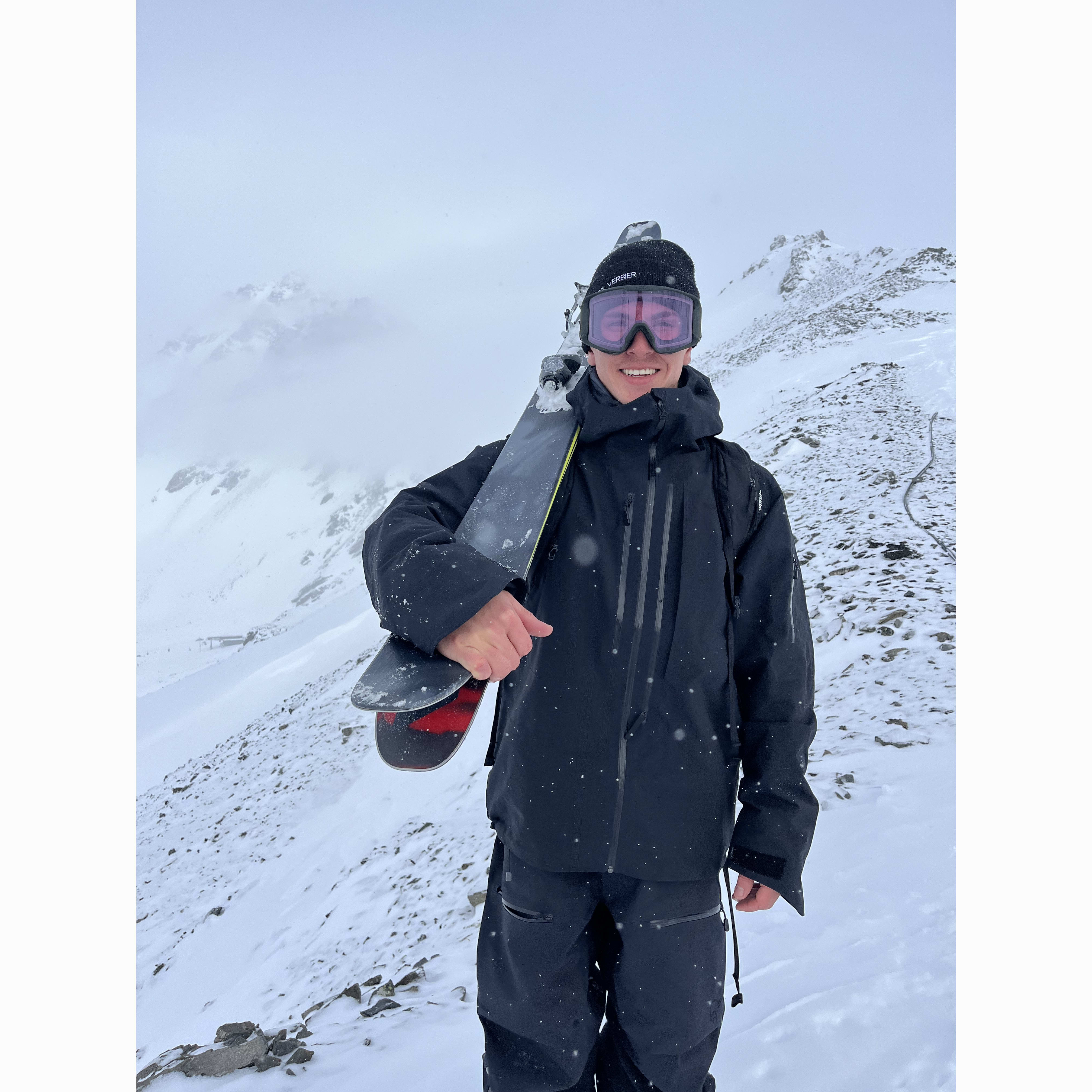 A smiling skier in a black Norrøna jacket and goggles, holding skis on a snowy mountain.