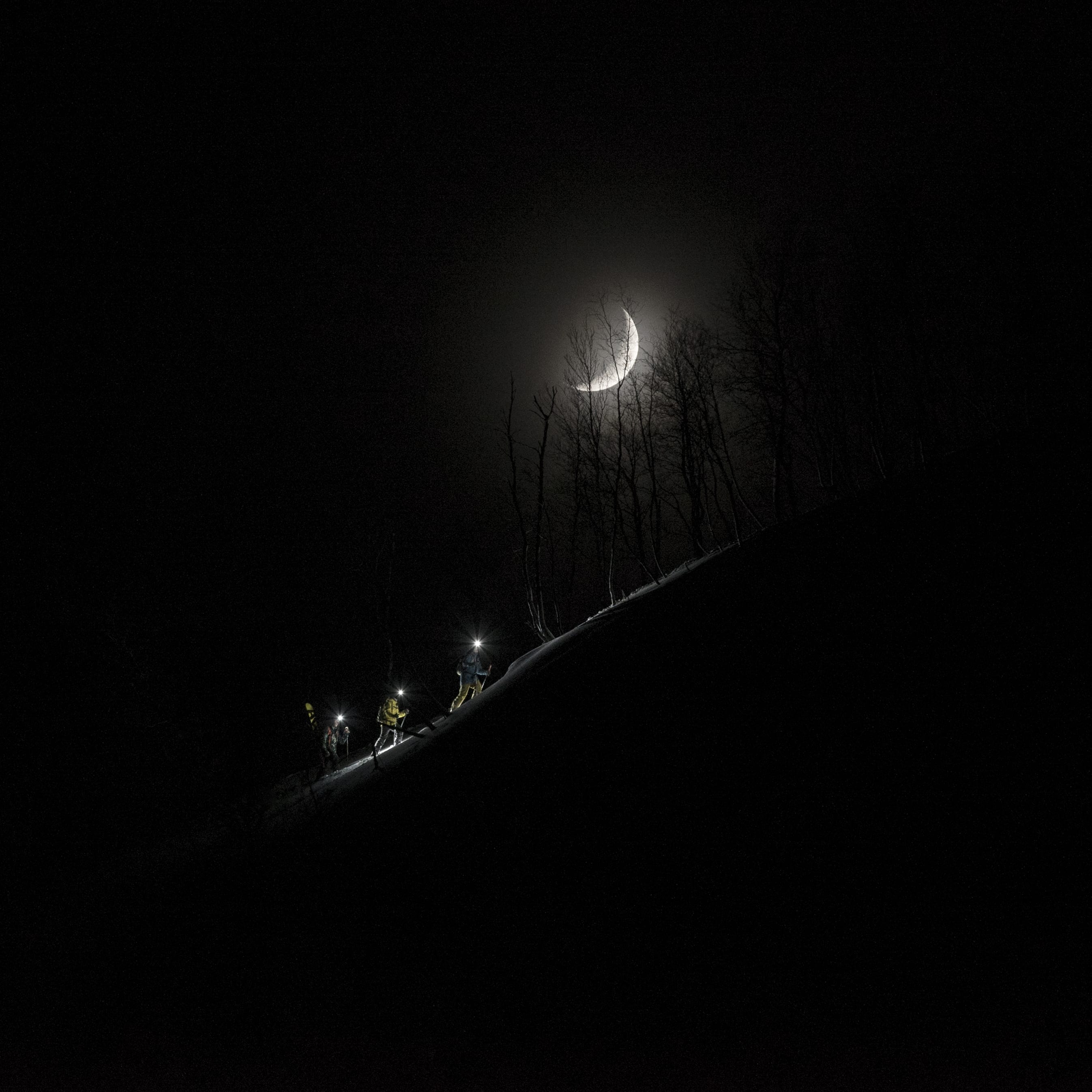 Three people wearing headlamps walk up a snowy hill with a crescent moon in the sky.