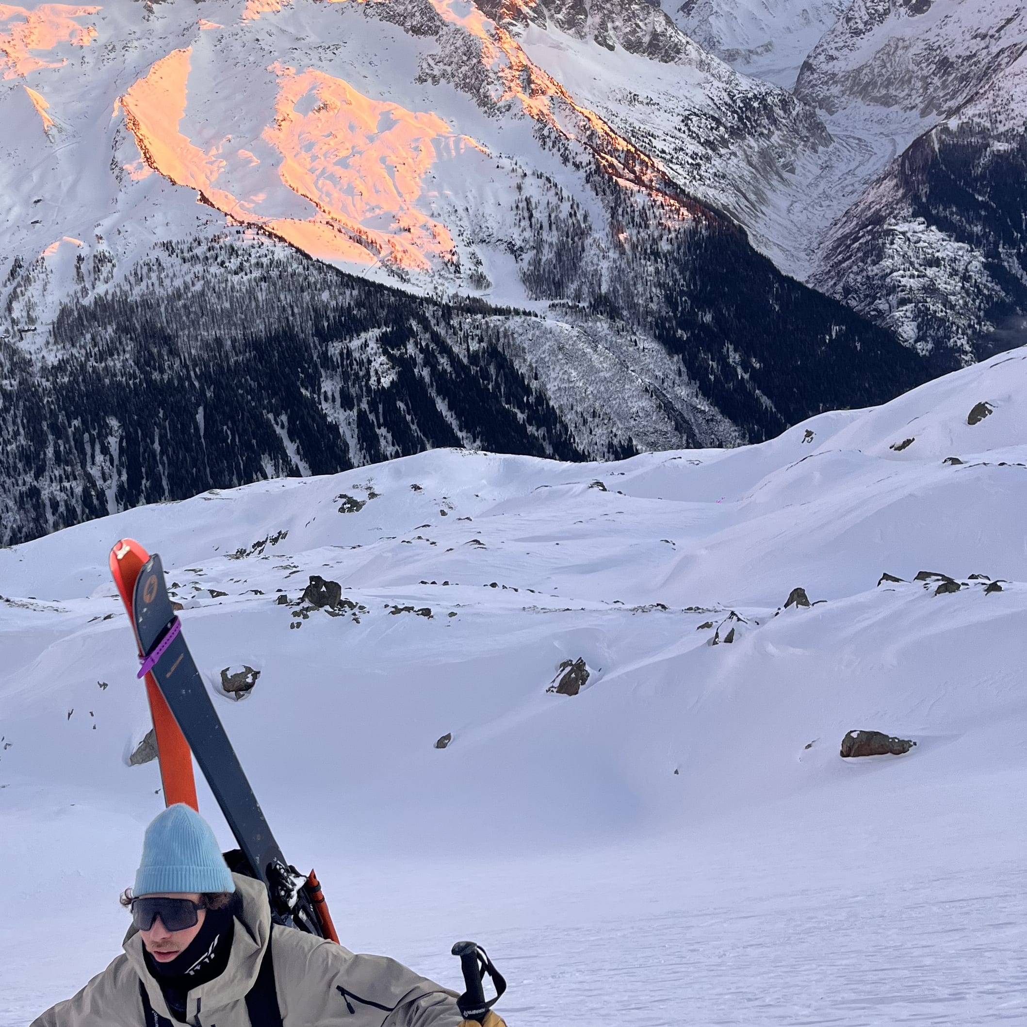 A person in a Norrøna ski suit is skiing down a snowy mountain slope. The sun is setting in the background, casting a warm glow on the mountain. 
