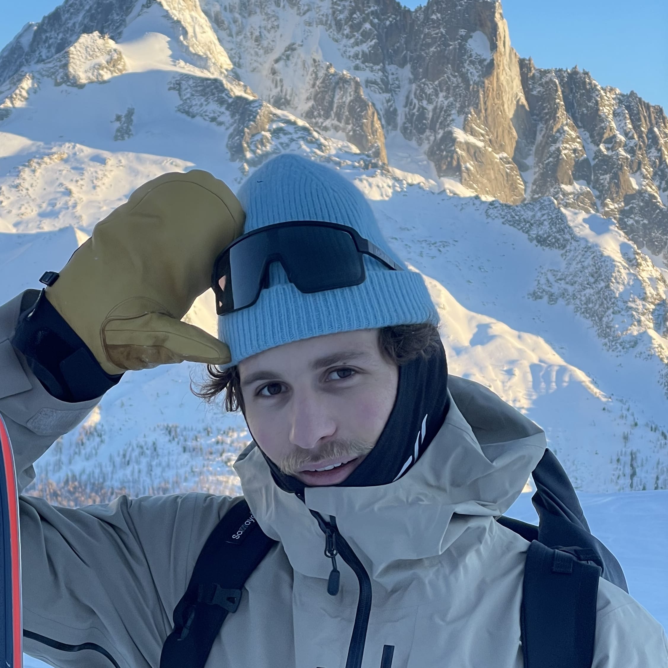 A person in a Norrøna jacket and a blue beanie holds a Blizzard ski in front of a snowy mountain range.