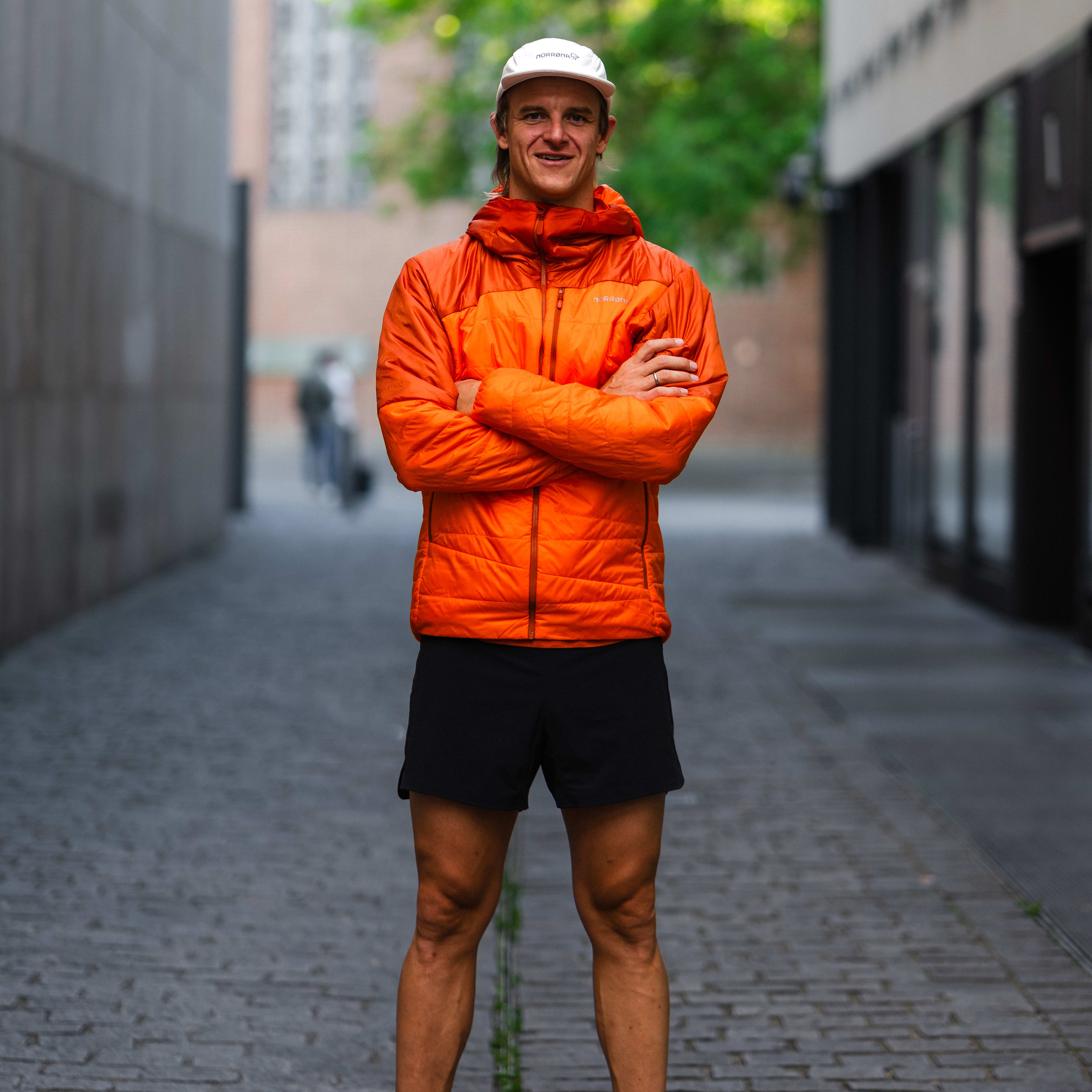A man stands in an alleyway, arms crossed, wearing a Norrøna jacket, shorts, and a cap.