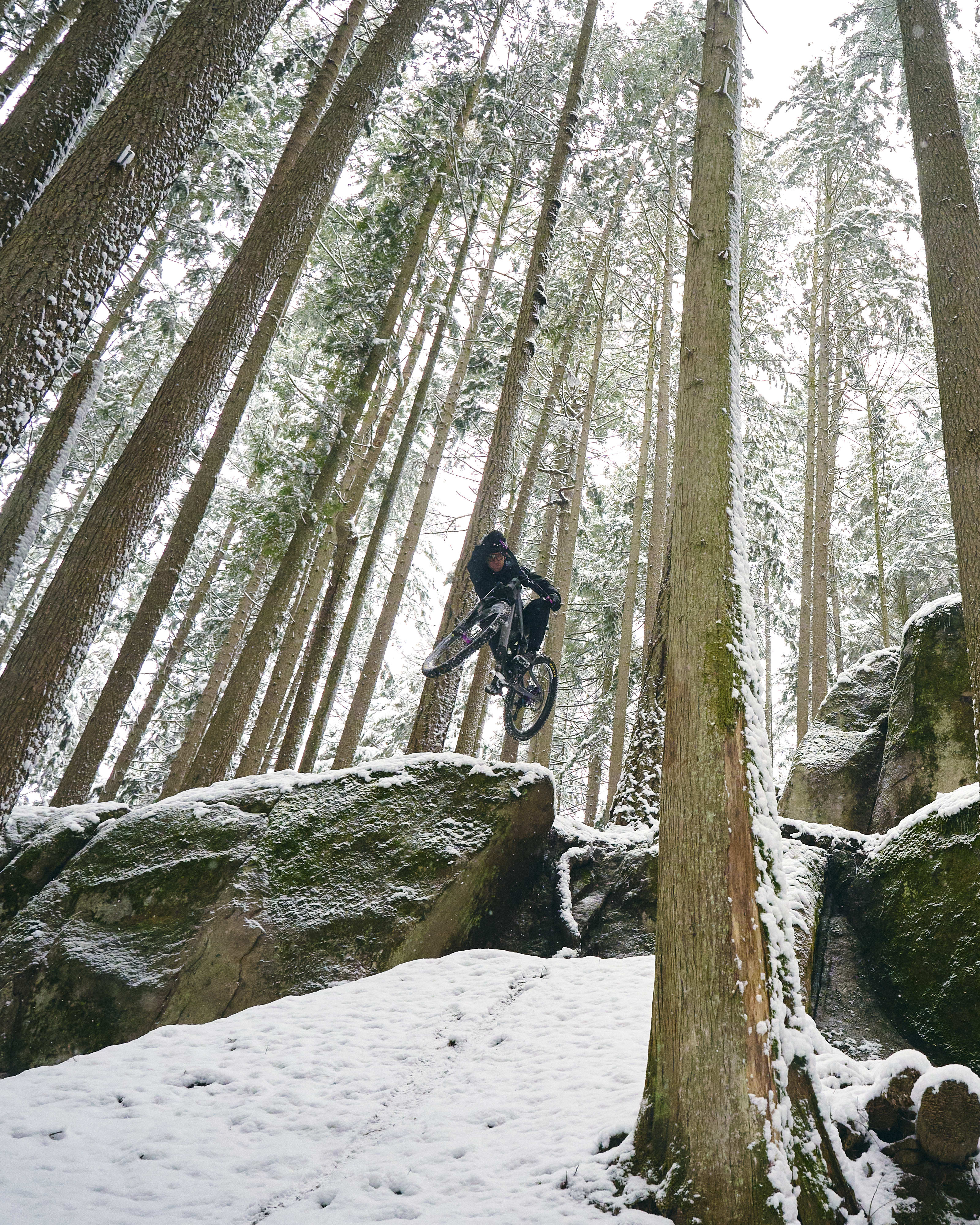 A person wearing Norrøna clothing mountain bikes through a snowy forest. The biker is airborne over a large rock. The background is a snow-covered forest.