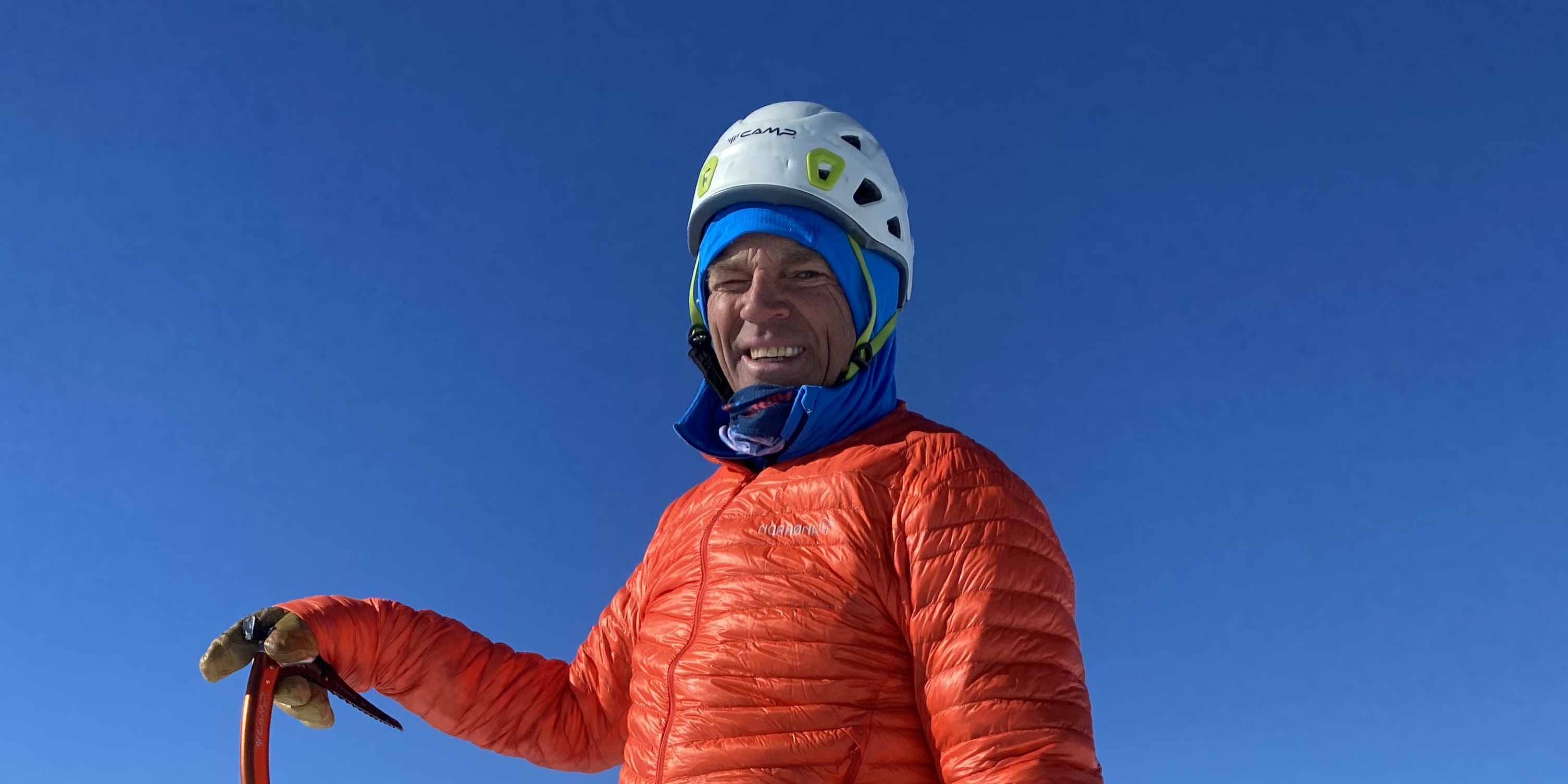 A man wearing a Norrøna jacket and pants, a helmet, and ice climbing gear stands on a snowy mountain peak. 
