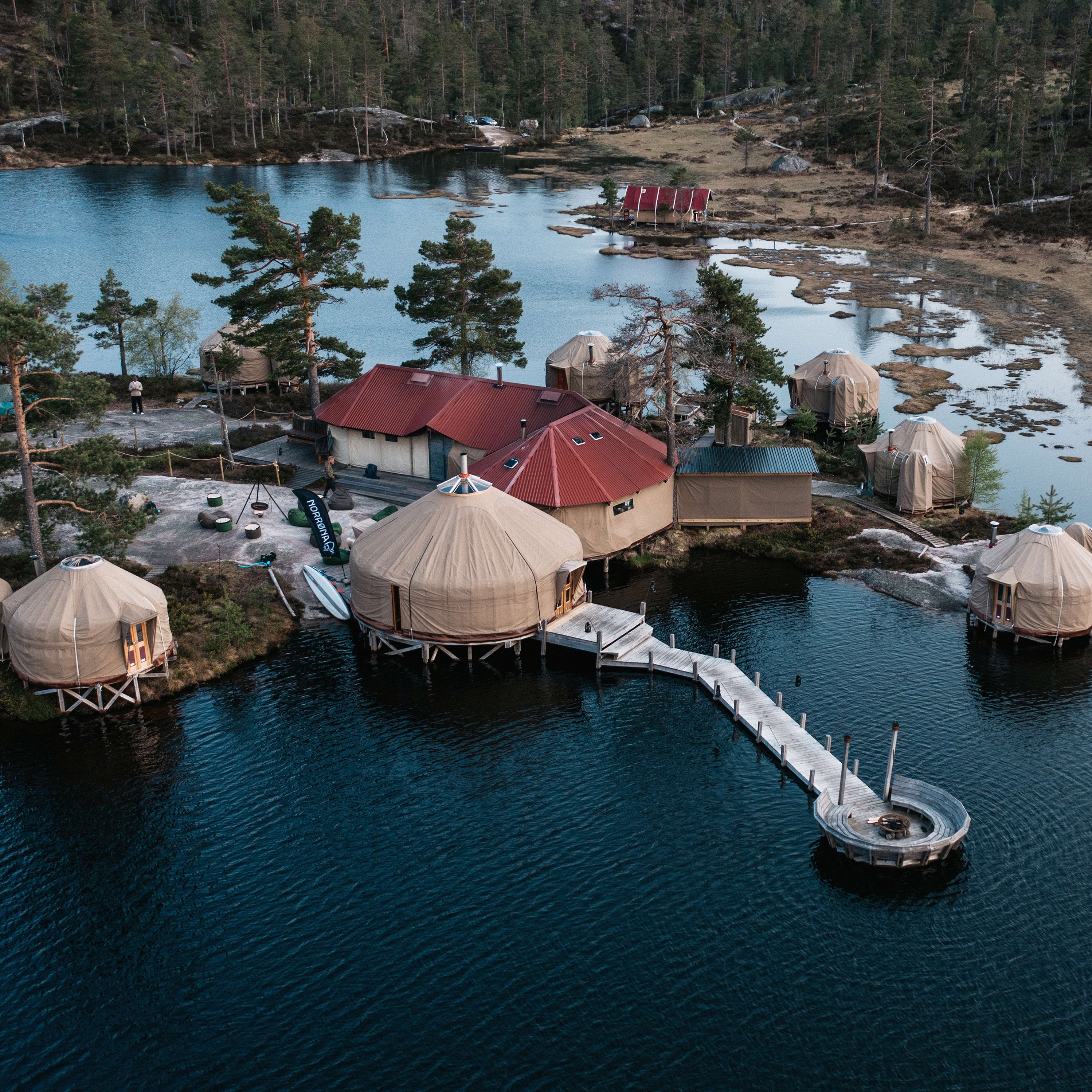 Aerial view of canvas yurts connected by boardwalks on a lake with trees and a Norrøna banner.