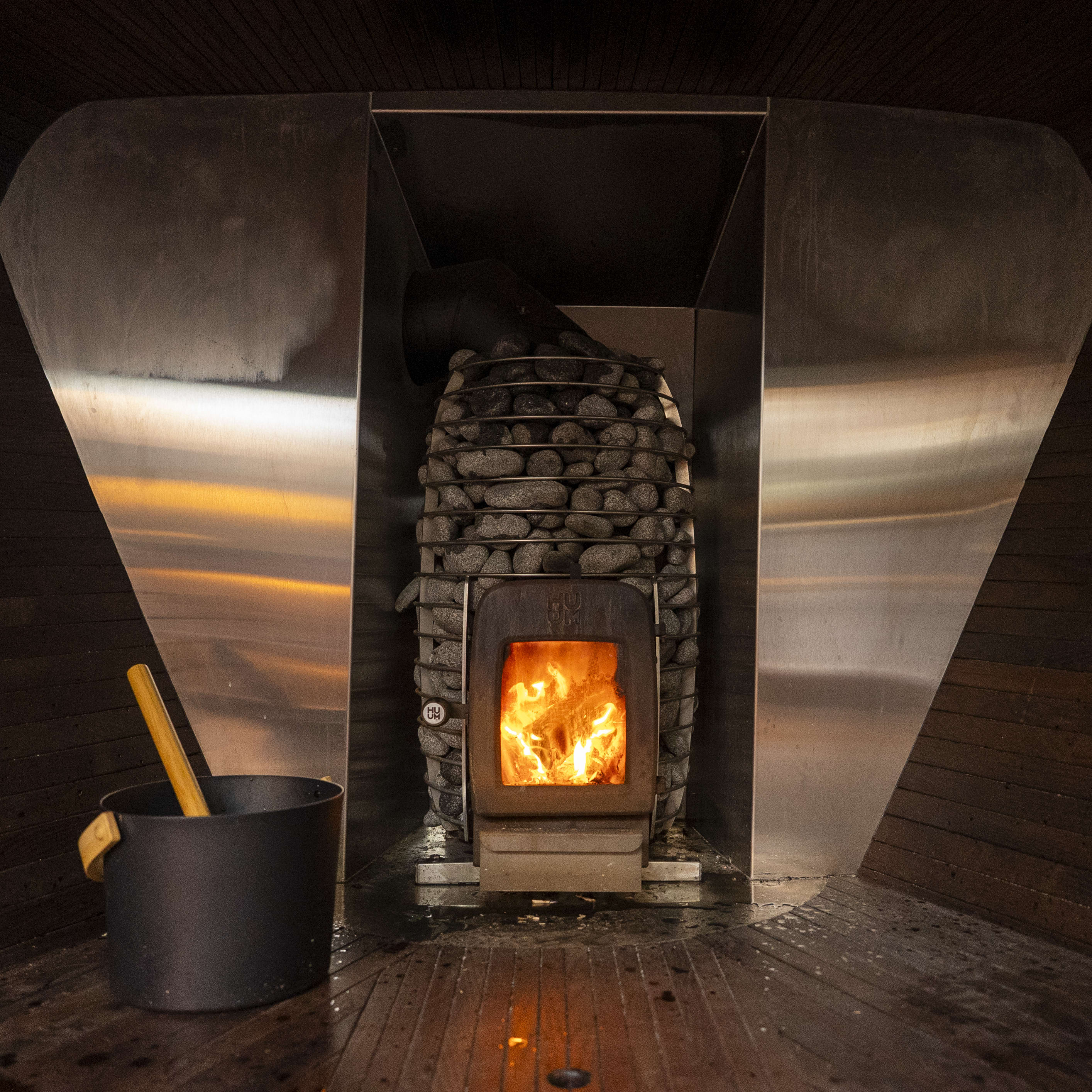 Sauna interior with wood-burning stove and bucket with a ladle. Visible flames inside the stove.