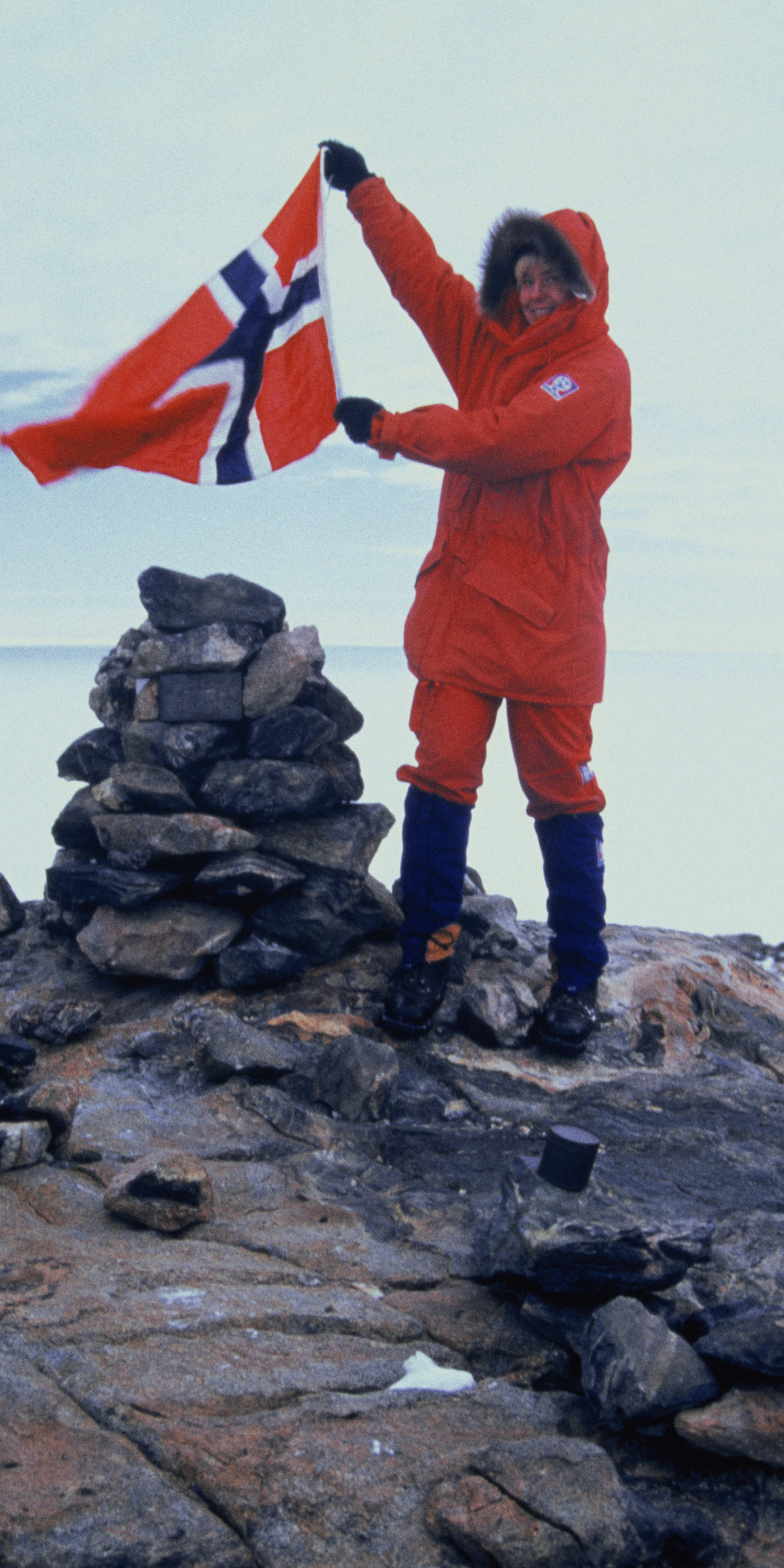 A person in a red jacket and blue pants stands on rocky terrain holding a waving Norwegian flag.  They stand near a small cairn of rocks.