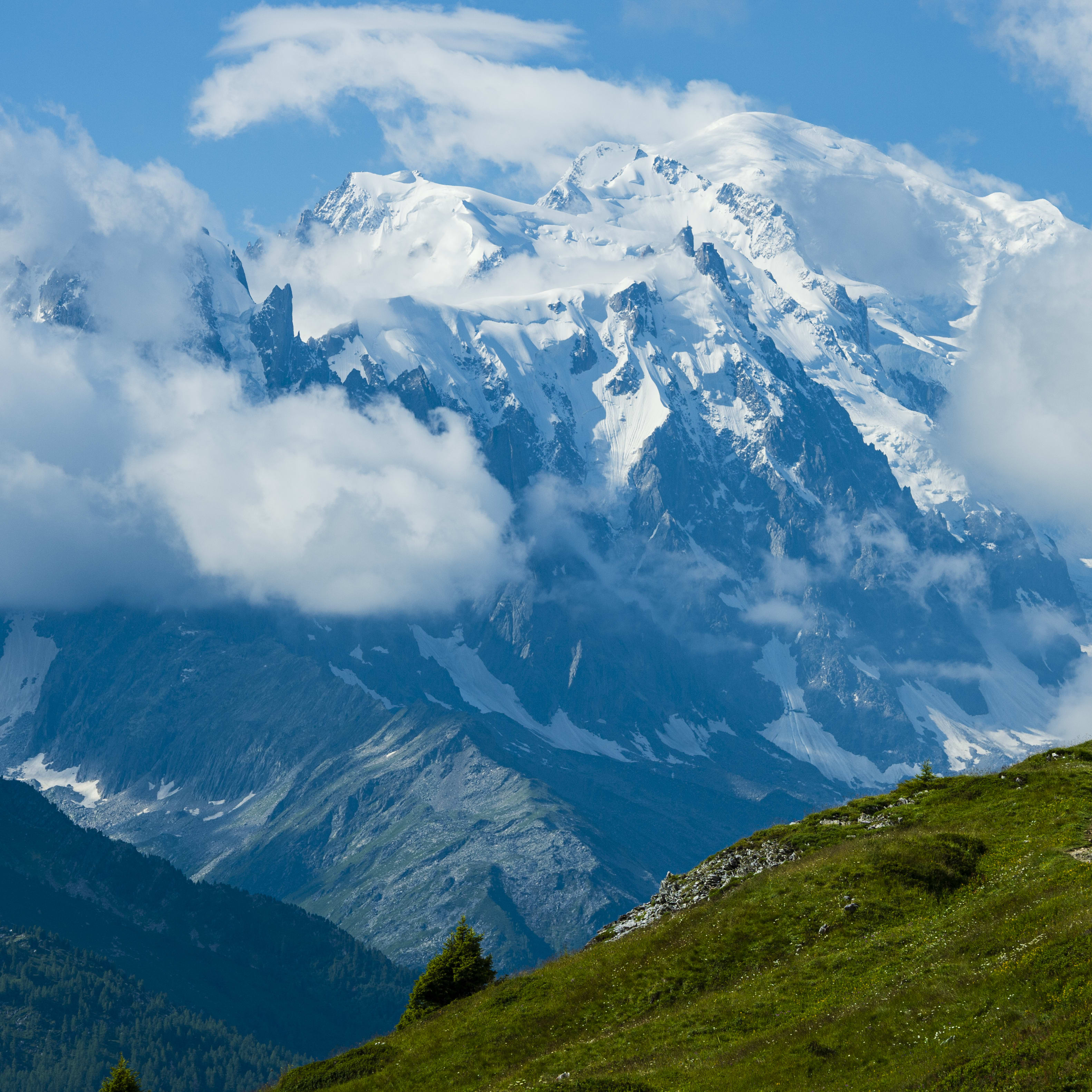 A person is biking on a trail, going uphill, towards a large snow-capped mountain. The mountain is partially obscured by clouds. The person is wearing Norrøna clothing. 

