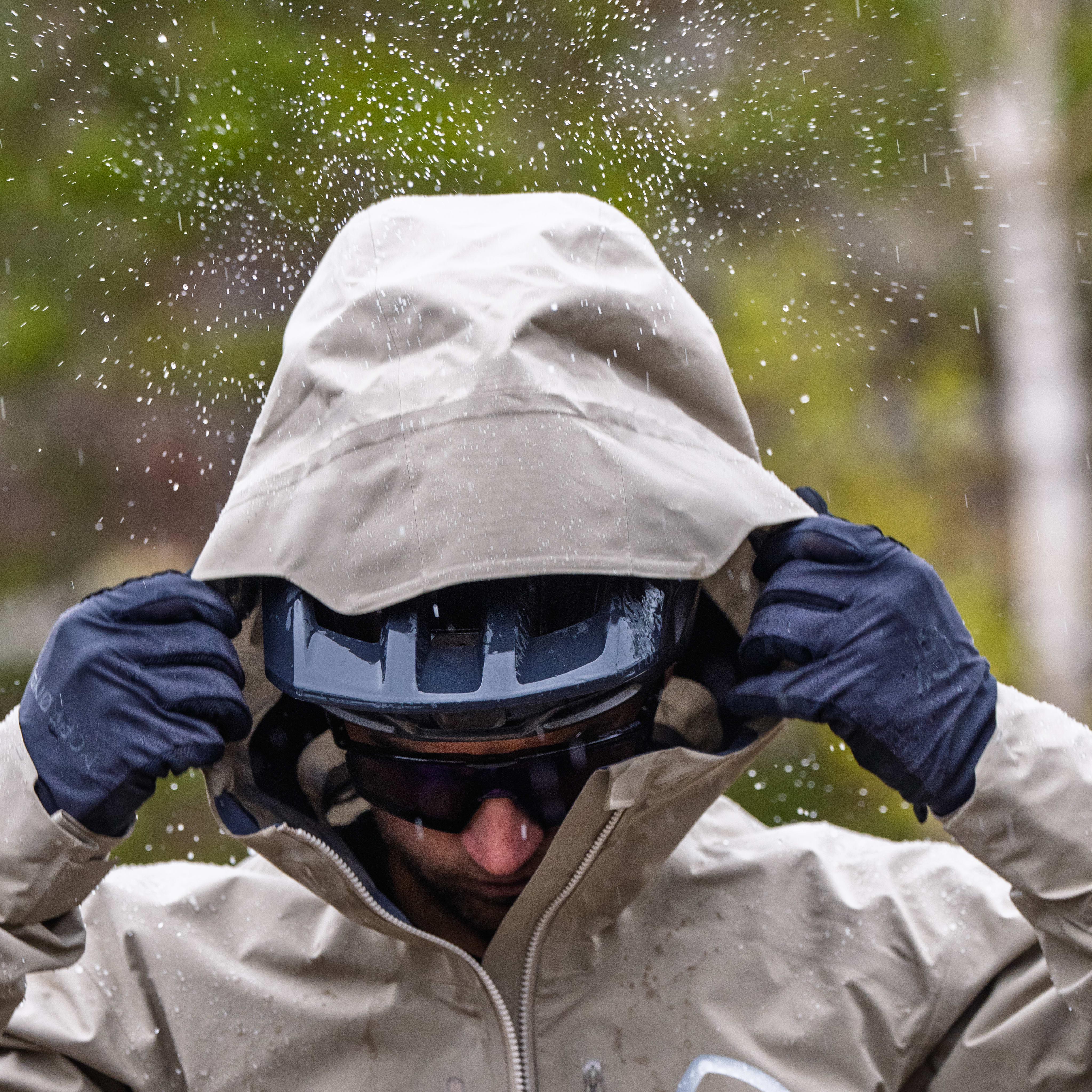 A cyclist pulls the hood of a beige Norrøna jacket over their helmet in the rain.